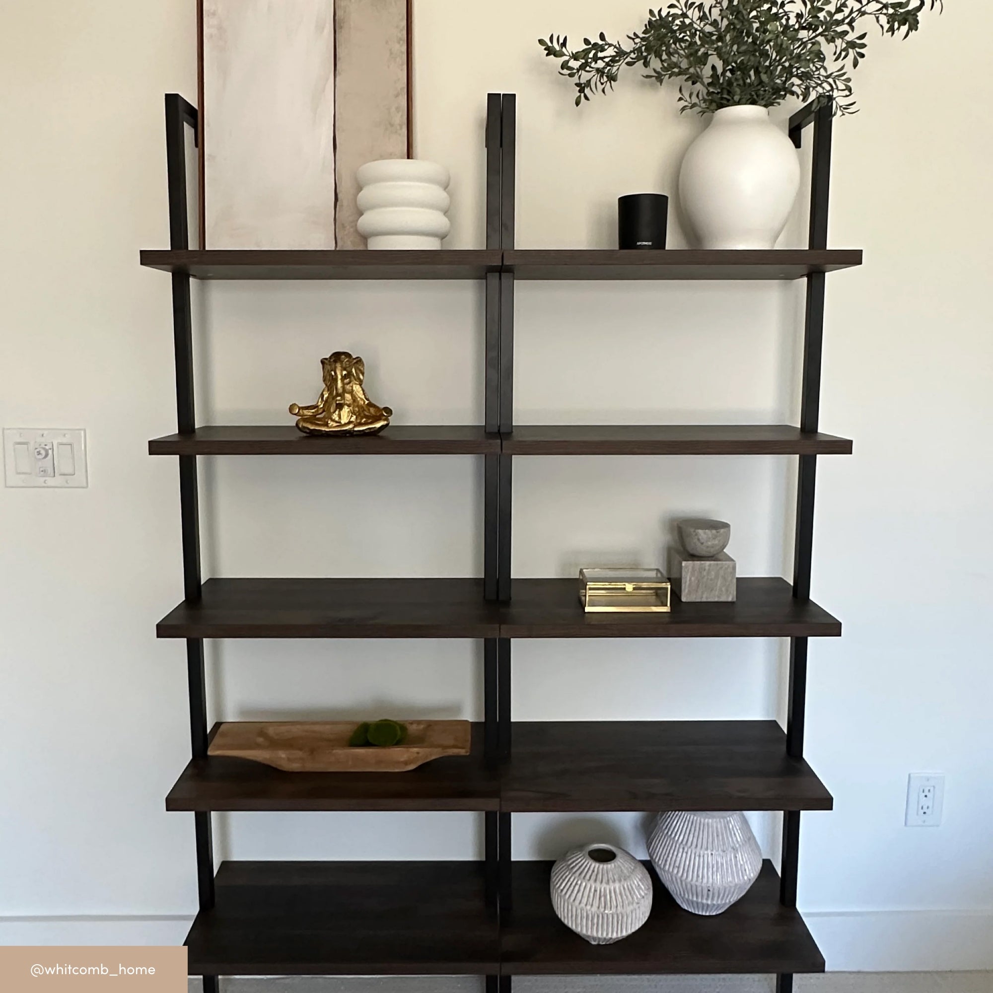 A modern wooden shelving unit with black metal supports displays decorative items, including vases, stacked bowls, a gold sculpture, a tray with green objects, a plant, and framed artwork against a white wall.