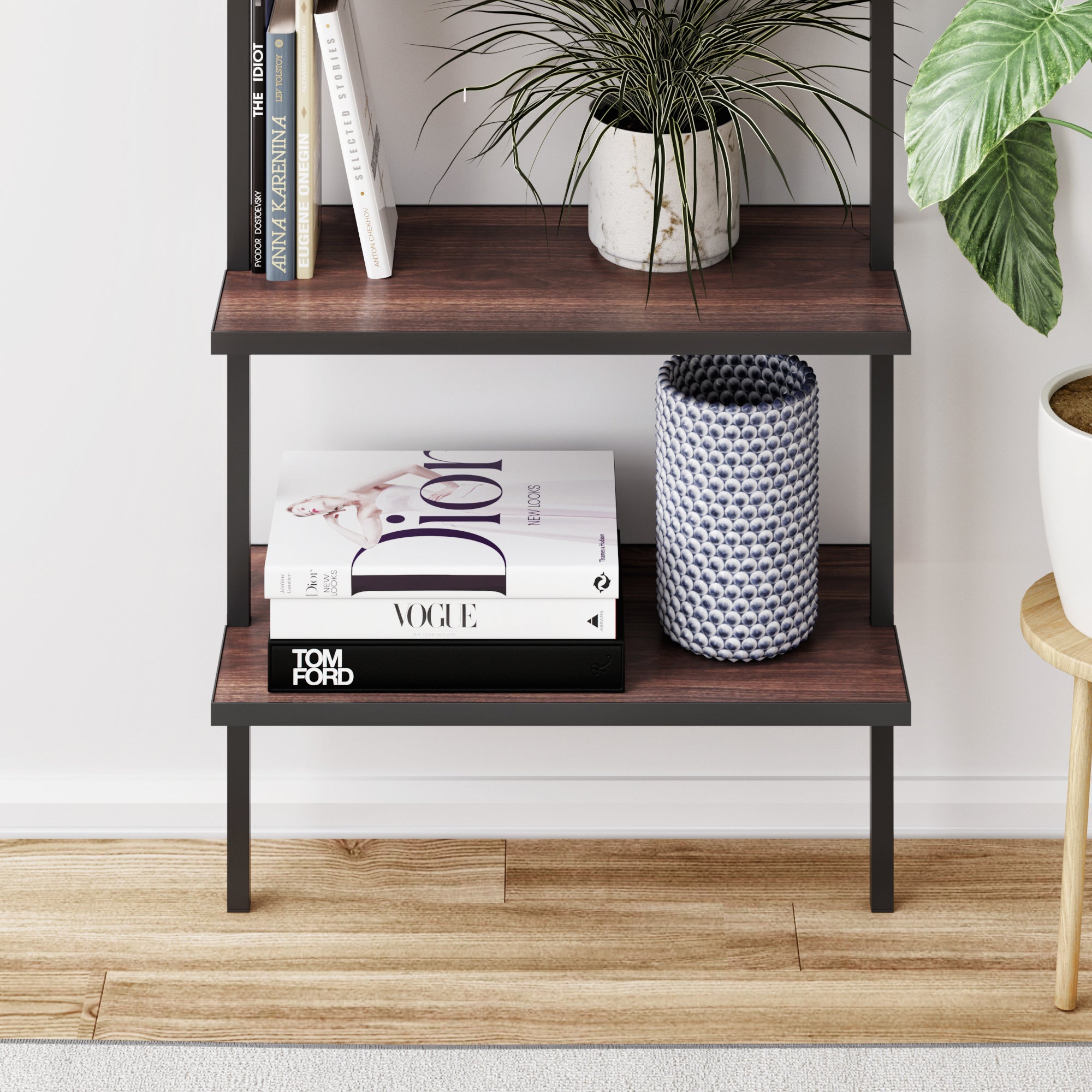 A bookshelf with wood shelves holds books, a potted plant, and a blue-patterned vase. Visible book titles include “Dior” and “Vogue.” The bookshelf stands on a wooden floor next to a small stool.
