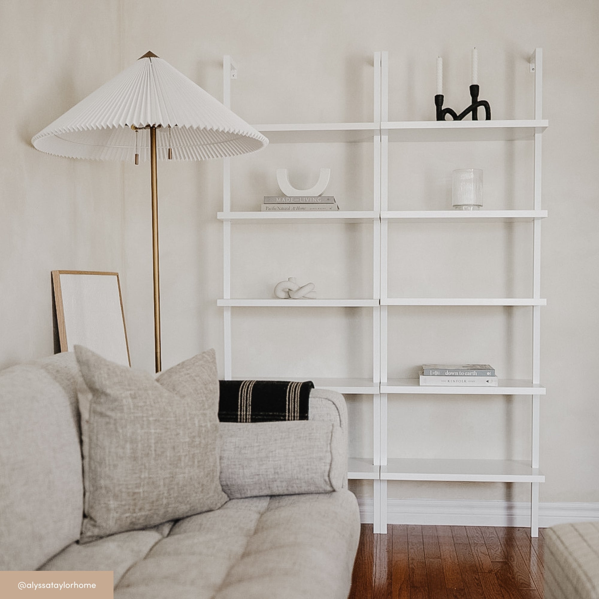 Minimalist living room with a beige sofa, textured pillows, a modern white bookshelf with a few decorative objects, a sculptural black candle holder, and a white floor lamp with a pleated shade on a wooden floor.