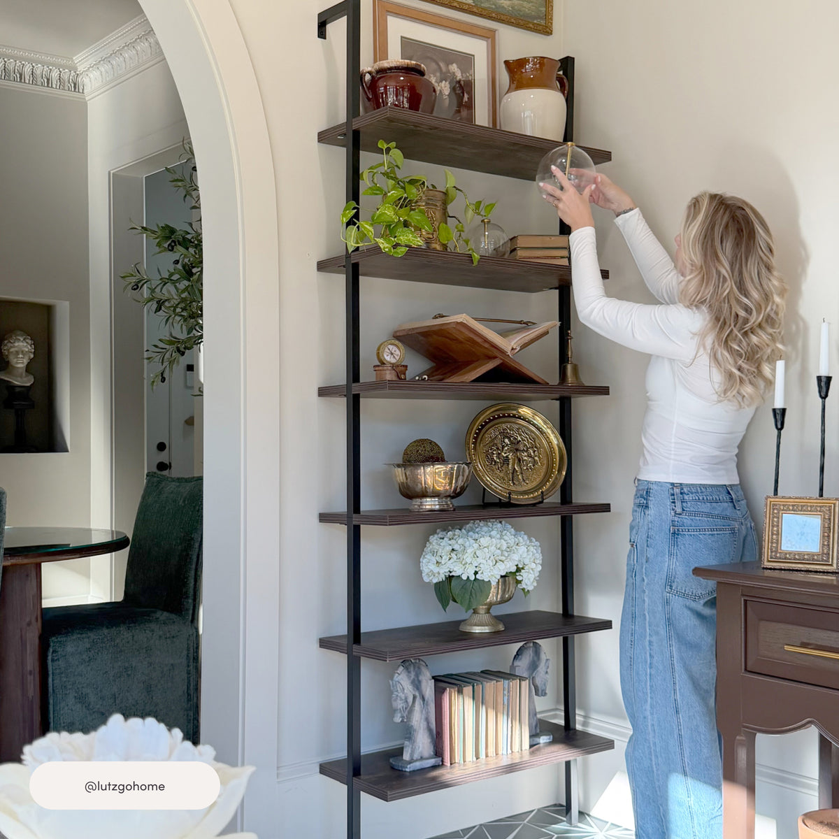 A woman places a decorative glass sphere on the top shelf of a Nathan James 6-Shelf Ladder Bookshelf Nutmeg Matte Black, which is filled with books and home decor items.