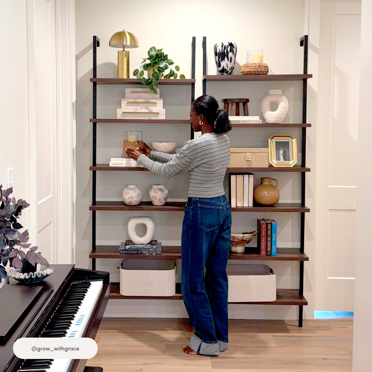 A person stands in front of a Nathan James 6-Shelf Ladder Bookshelf Nutmeg Matte Black, adjusting a decorative item on one of its six sturdy shelves.