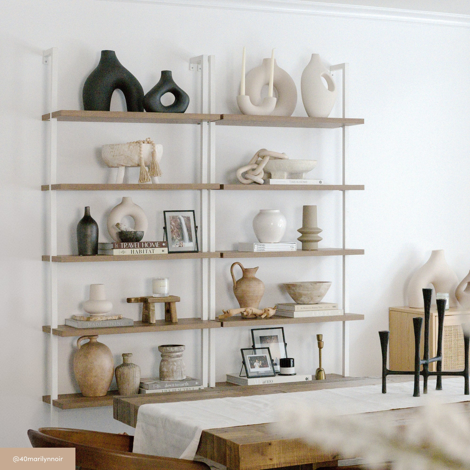 A modern wooden wall shelf displays neutral-toned ceramic vases, bowls, books, framed photos, and decorative objects in various shapes, arranged neatly in a minimalist dining room with a wooden table below.