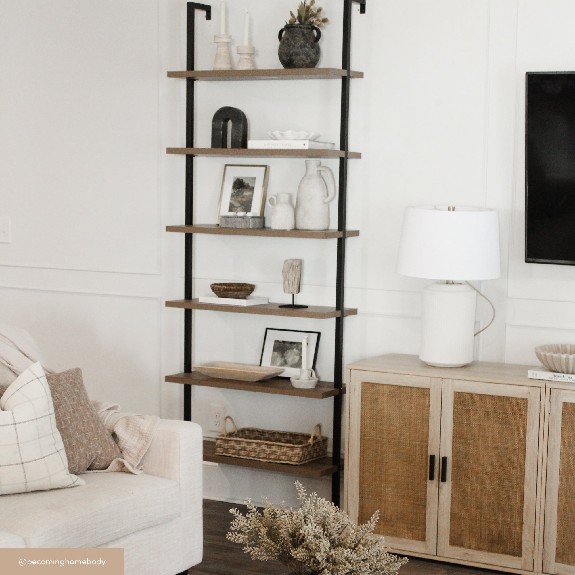 A tall, black-framed shelving unit with wooden shelves displays vases, framed art, books, and decor beside a white and rattan cabinet, a white lamp, and a beige couch in a neutral living room.
