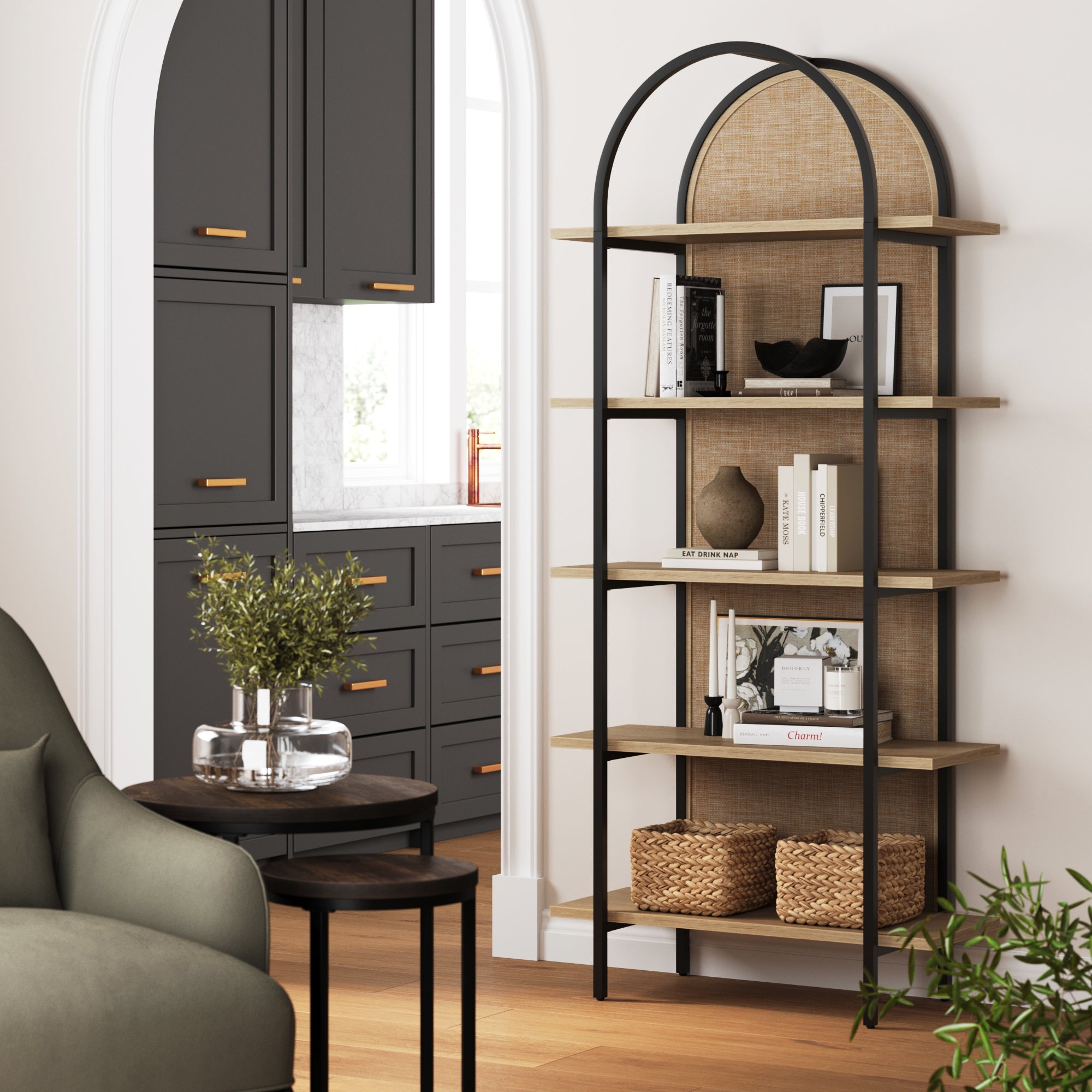 A modern living space featuring a black and wood arched bookshelf with books, decor, and baskets. Nearby are a green armchair, round tables with plants, and a view into a kitchen with gray cabinets and gold hardware.