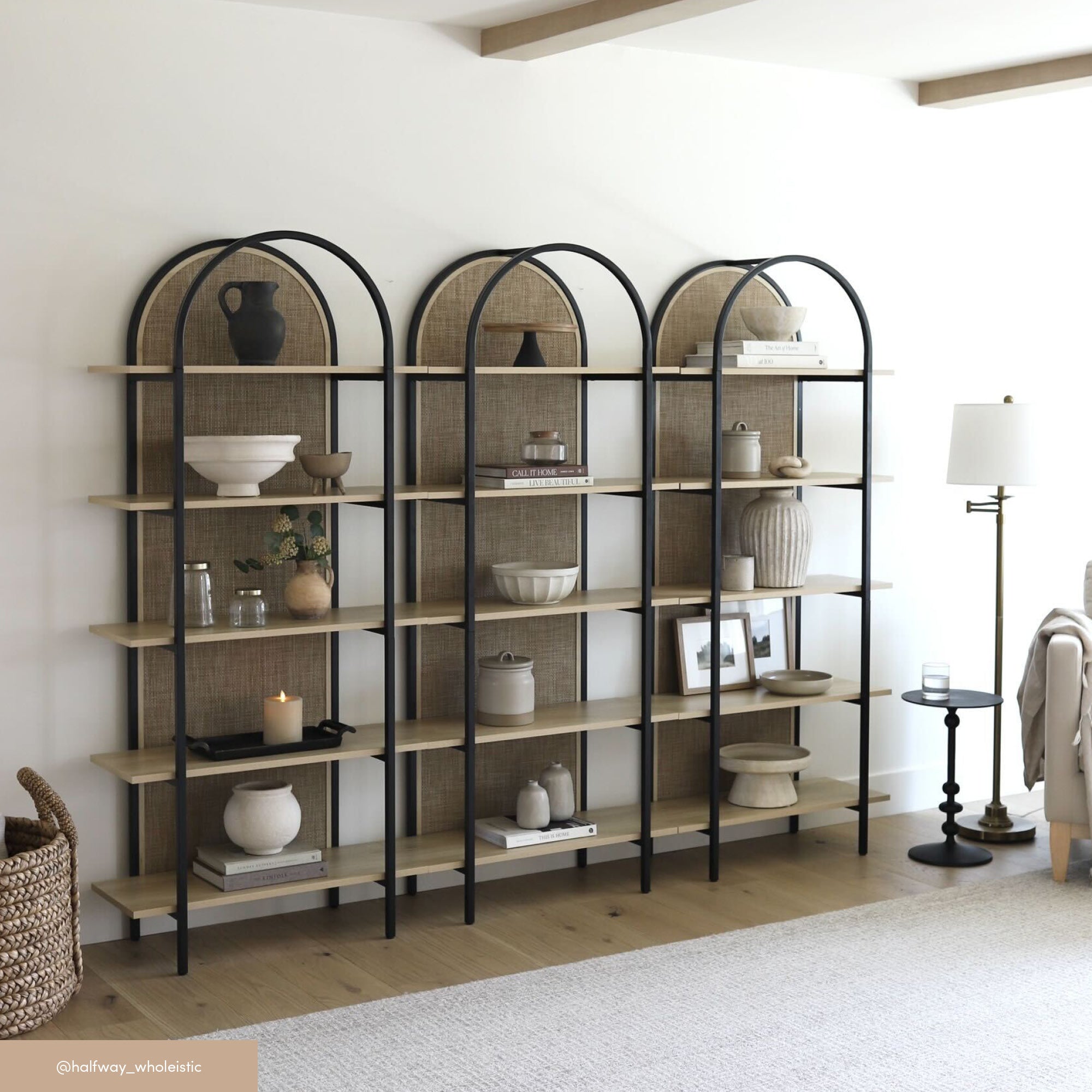 Three tall, black-framed bookshelves with arched tops hold neutral-toned decorative items, pottery, and framed photos. A white floor lamp and beige armchair are nearby in a modern, minimalist living room.