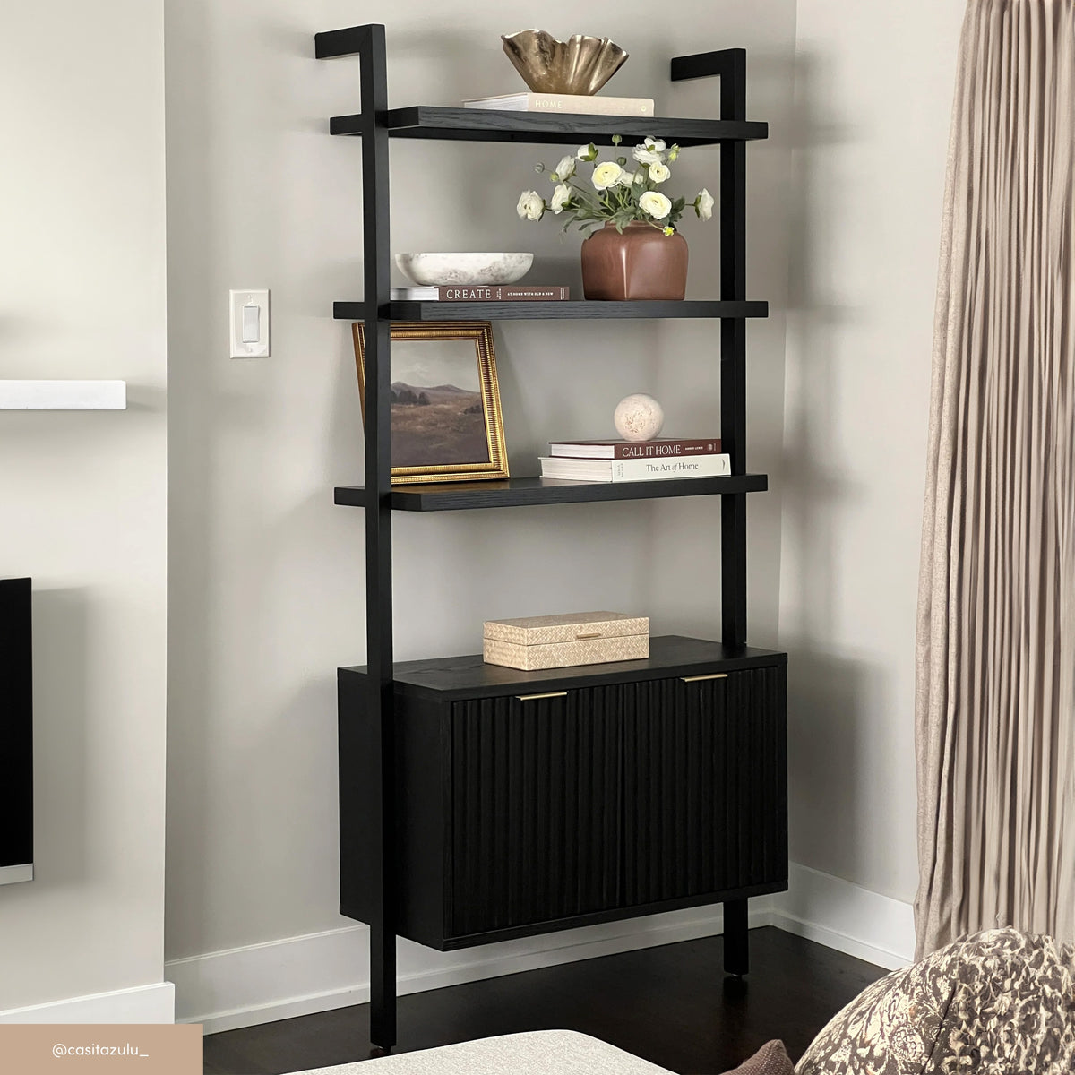 A modern black shelving unit with three open shelves displaying a bowl, a vase of white flowers, books, a framed photo, and decorative objects, stands against a light-colored wall in a living room.