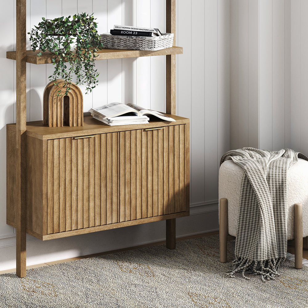 A light wood wall-mounted cabinet with two slatted doors, topped with decor and books, next to a small upholstered stool draped with a patterned throw blanket.