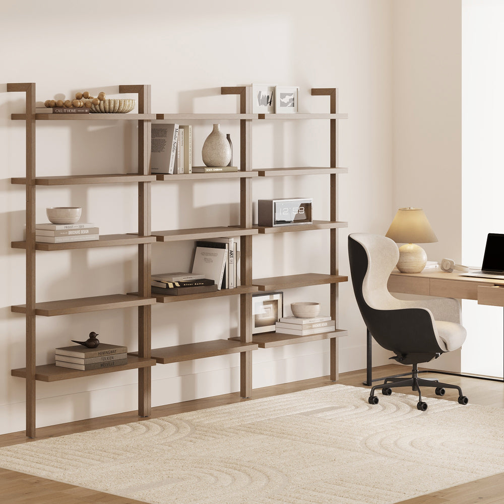 A wall-mounted modular wooden shelving unit displays books, decorative objects, and a digital clock, positioned next to a desk with an office chair and lamp.
