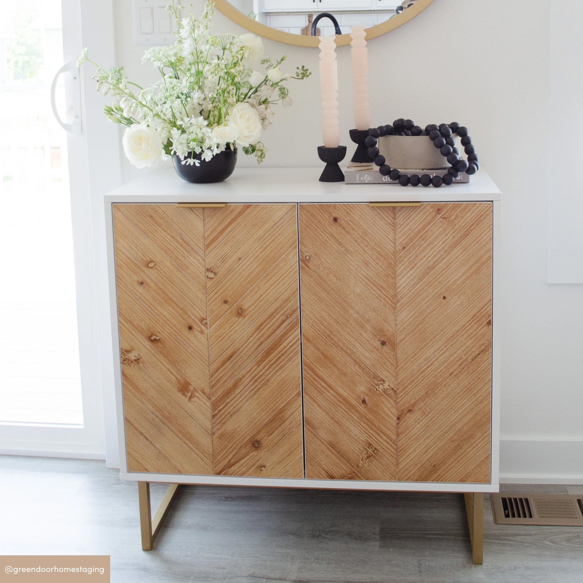 A modern sideboard with a white frame and chevron-patterned wooden doors stands against a white wall. On top are a floral arrangement, two stacked candles, black beads, and a round mirror hanging above.