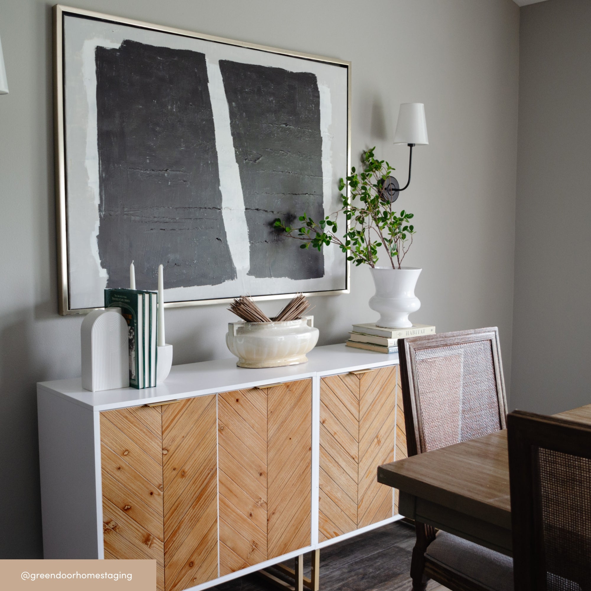 A modern dining room features a white sideboard with wood chevron doors, decorative books, vases, and a leafy plant. Above it hangs a large abstract painting with dark gray shapes. Two cane-backed chairs sit nearby.