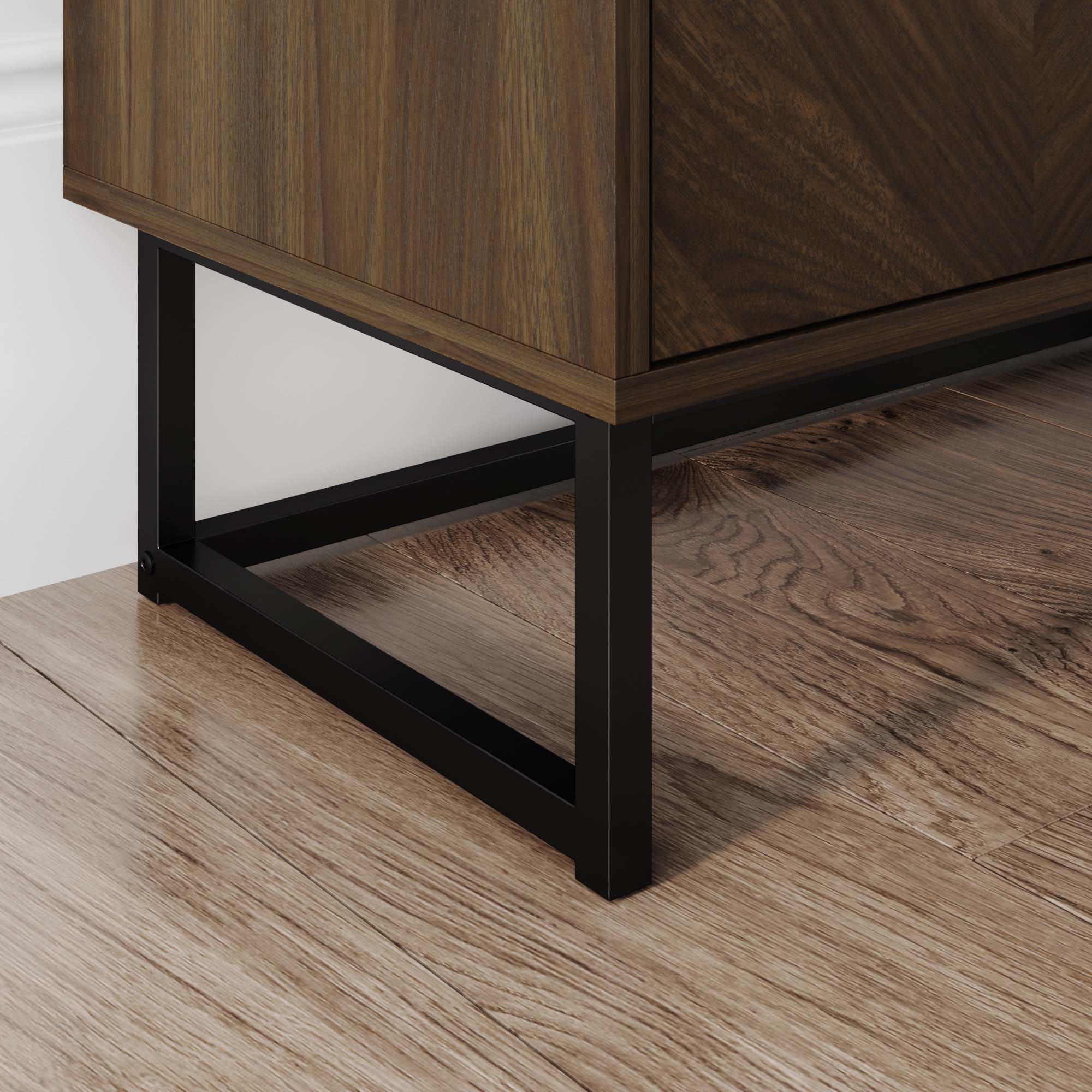 Close-up of the bottom corner of a wooden cabinet with a black metal frame, standing on light wood flooring next to a white wall.
