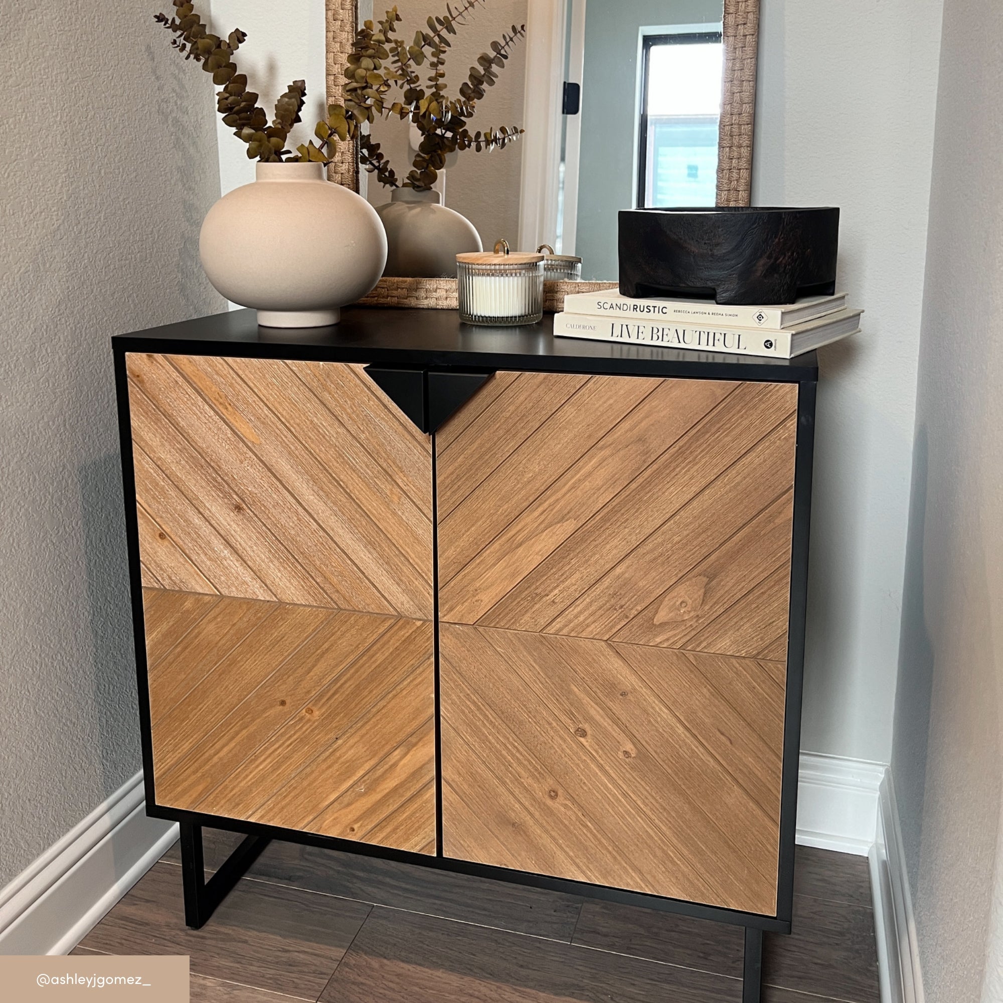A modern console table with chevron-patterned wooden doors stands against a light wall. On top are a ceramic vase with dried foliage, a candle, stacked books, and a black decorative bowl. A mirror hangs above.