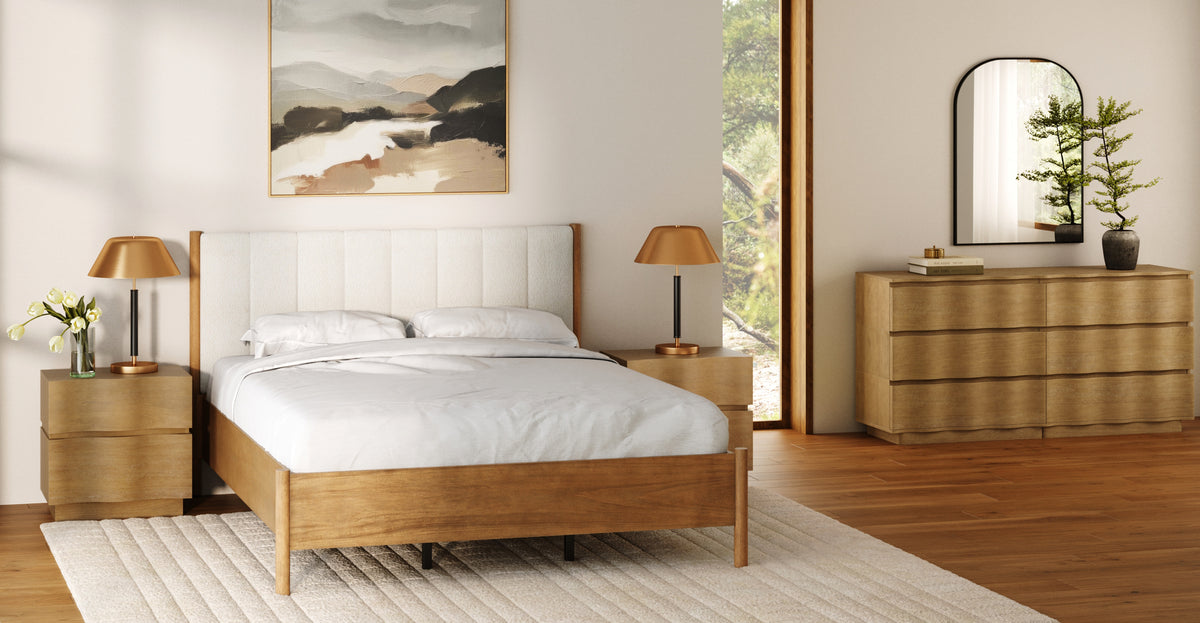 A modern bedroom featuring a bed with a light-colored upholstered headboard, wooden nightstands with gold lamps, and a matching dresser topped with a mirror and plant.