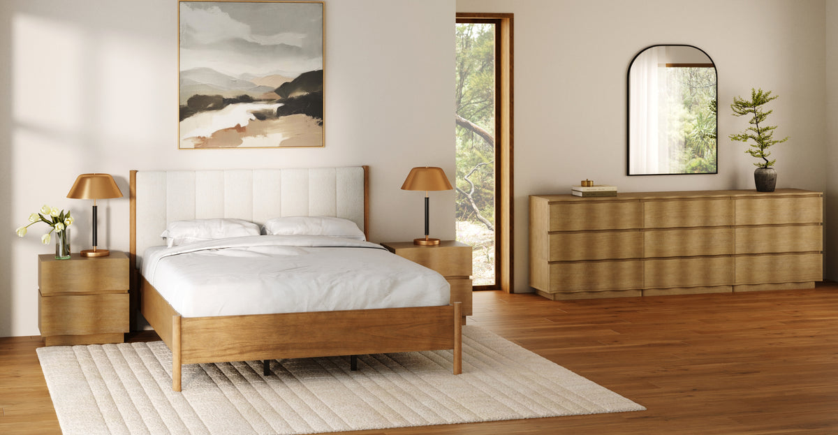 A bedroom with a wooden bed frame, two matching nightstands with gold lamps, a wooden dresser with a mirror, and an abstract landscape painting on the wall.