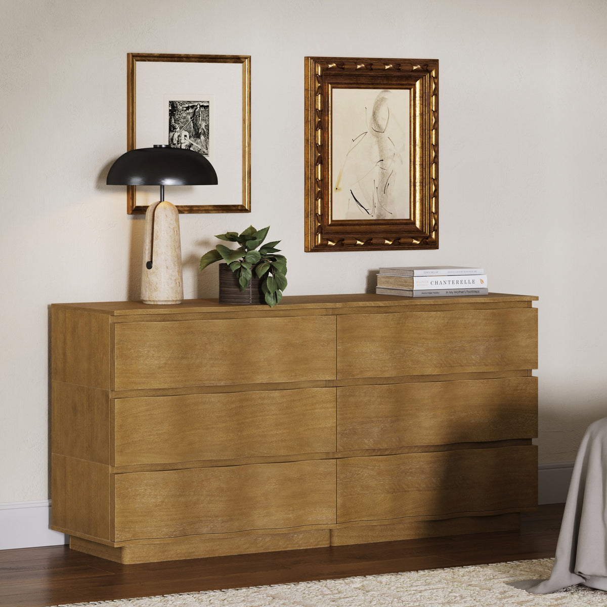 A light wood dresser with six drawers, topped by a table lamp, a small potted plant, and a stack of books, with two framed pictures hanging on the wall above.