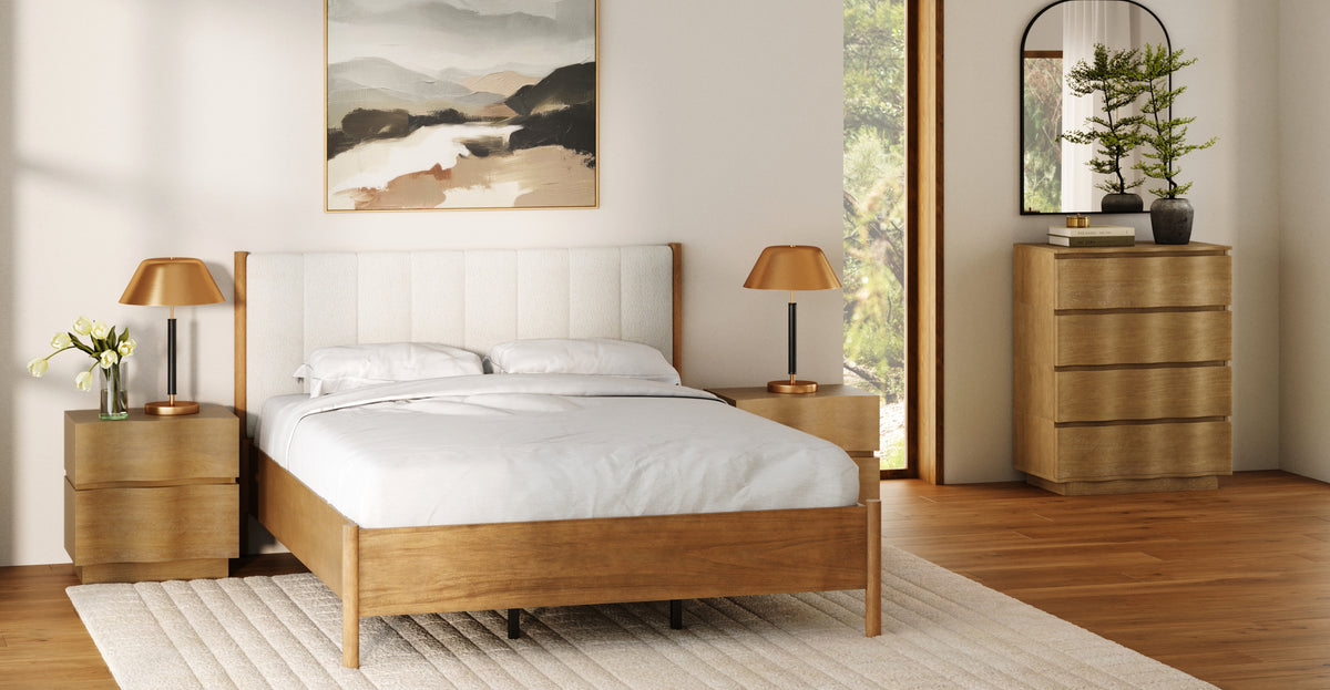 A bedroom featuring a wooden bed frame with a cream tufted headboard, two wooden nightstands with brass lamps, a matching wooden dresser, and a large abstract landscape painting.