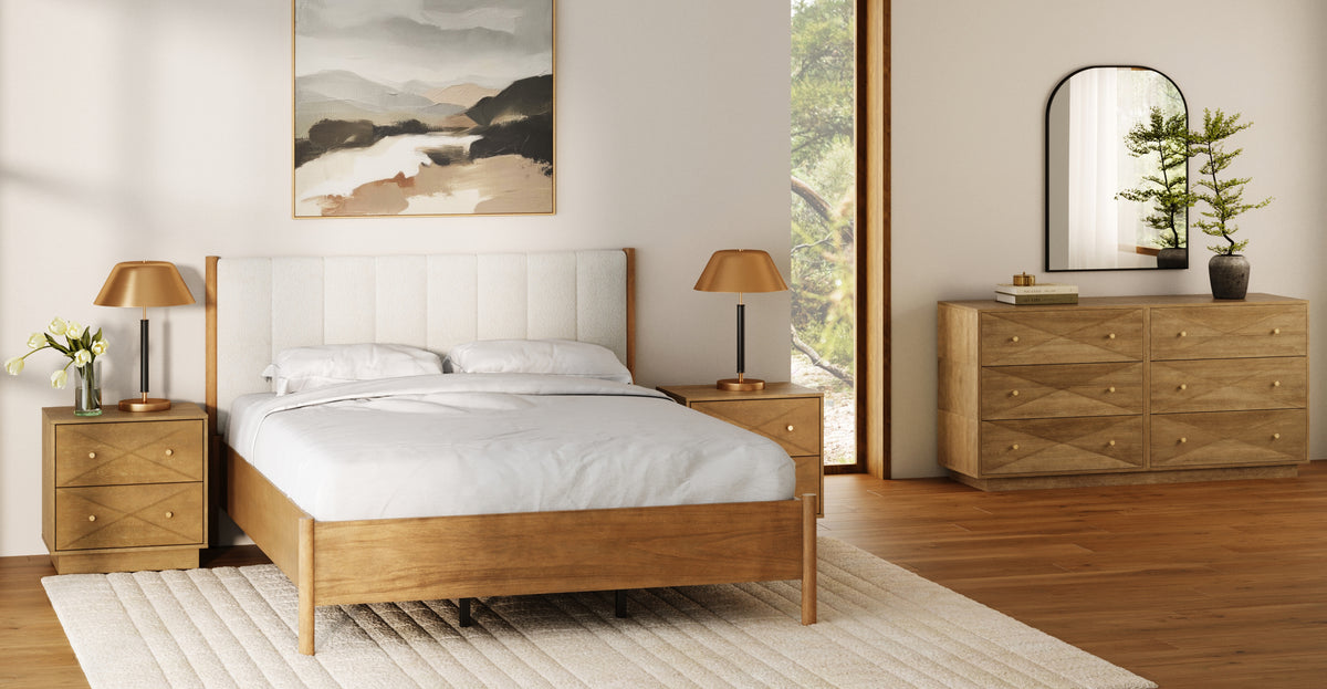 A bedroom with a wooden bed frame, two matching nightstands with brass-toned lamps, a six-drawer wooden dresser topped with a potted plant and mirror, and an abstract landscape painting on the wall.