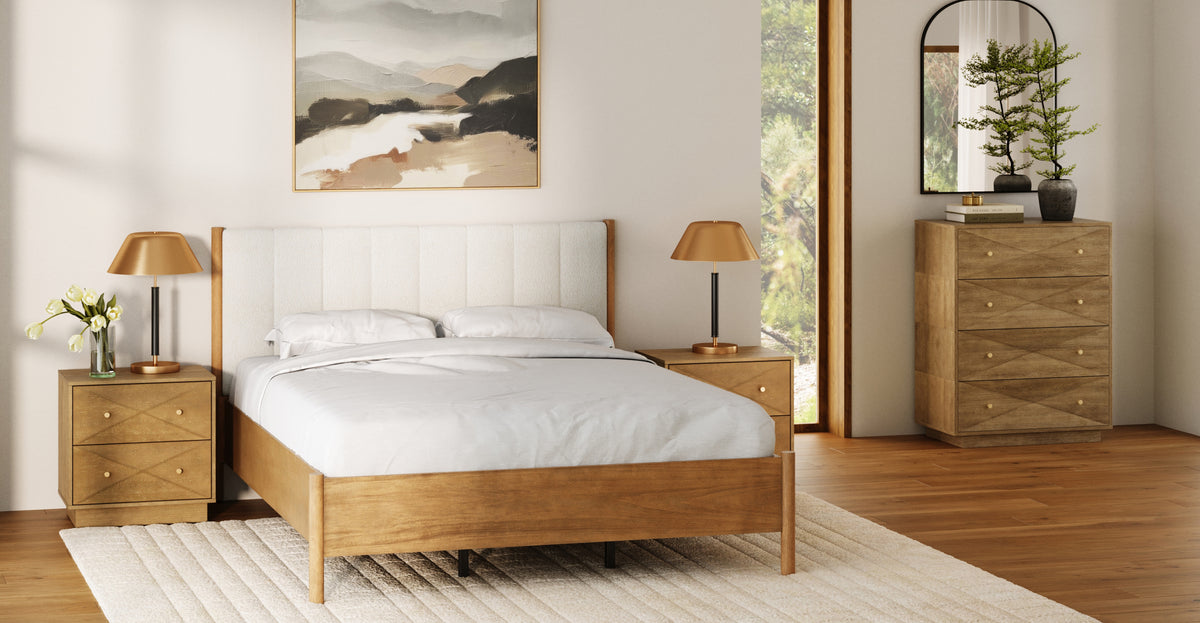 A wooden bed with a light-colored upholstered headboard, flanked by two matching wooden nightstands with gold lamps, and a tall dresser with a mirror, all set in a bedroom with a landscape painting on the wall.