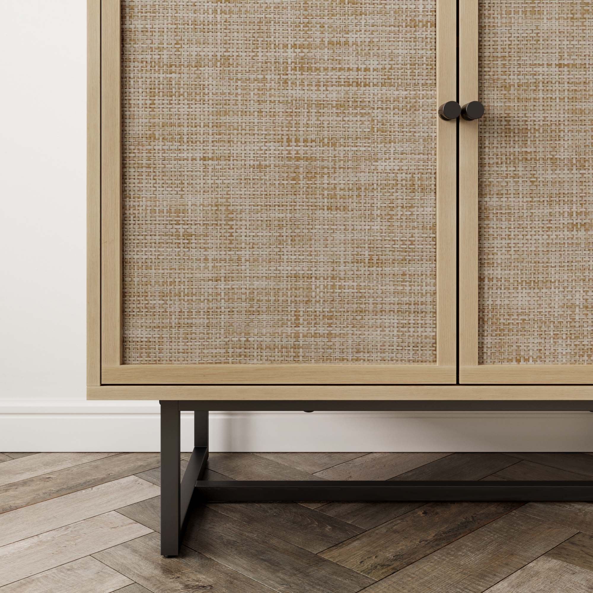 A close-up of a cabinet with woven rattan doors, light wood frame, round black knobs, and black metal legs, standing on a chevron-patterned wood floor against a white wall.