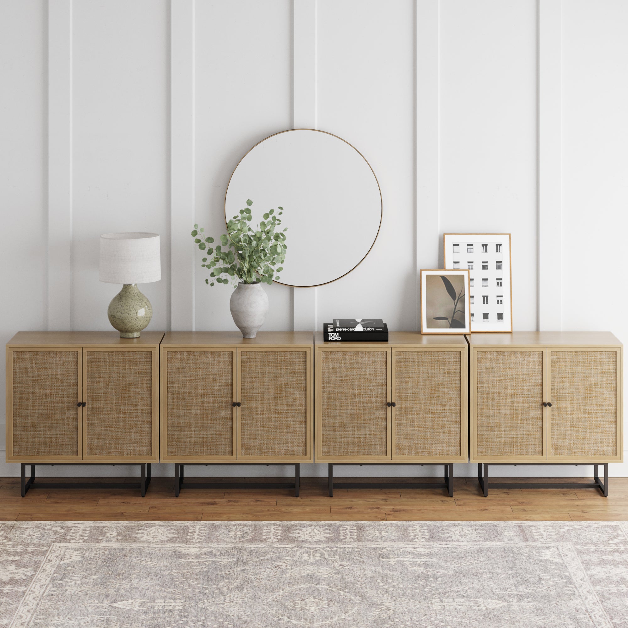A modern sideboard with four wooden cabinets sits against a white paneled wall. On top are a round mirror, a lamp, a vase with greenery, stacked books, and framed artwork. A patterned rug covers the wooden floor.