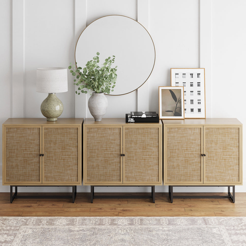 Three side-by-side wooden storage cabinets with cane-paneled doors, topped with a round mirror, table lamp, vase of greenery, and framed art.