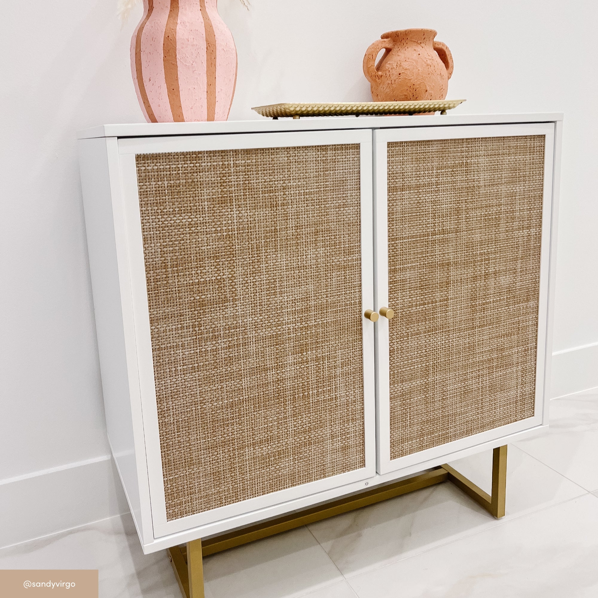 A white cabinet with textured, rattan-like doors stands on gold legs. On top are a striped pink vase, a terracotta jug, and a woven tray. The background features a white wall and light-colored floor tiles.