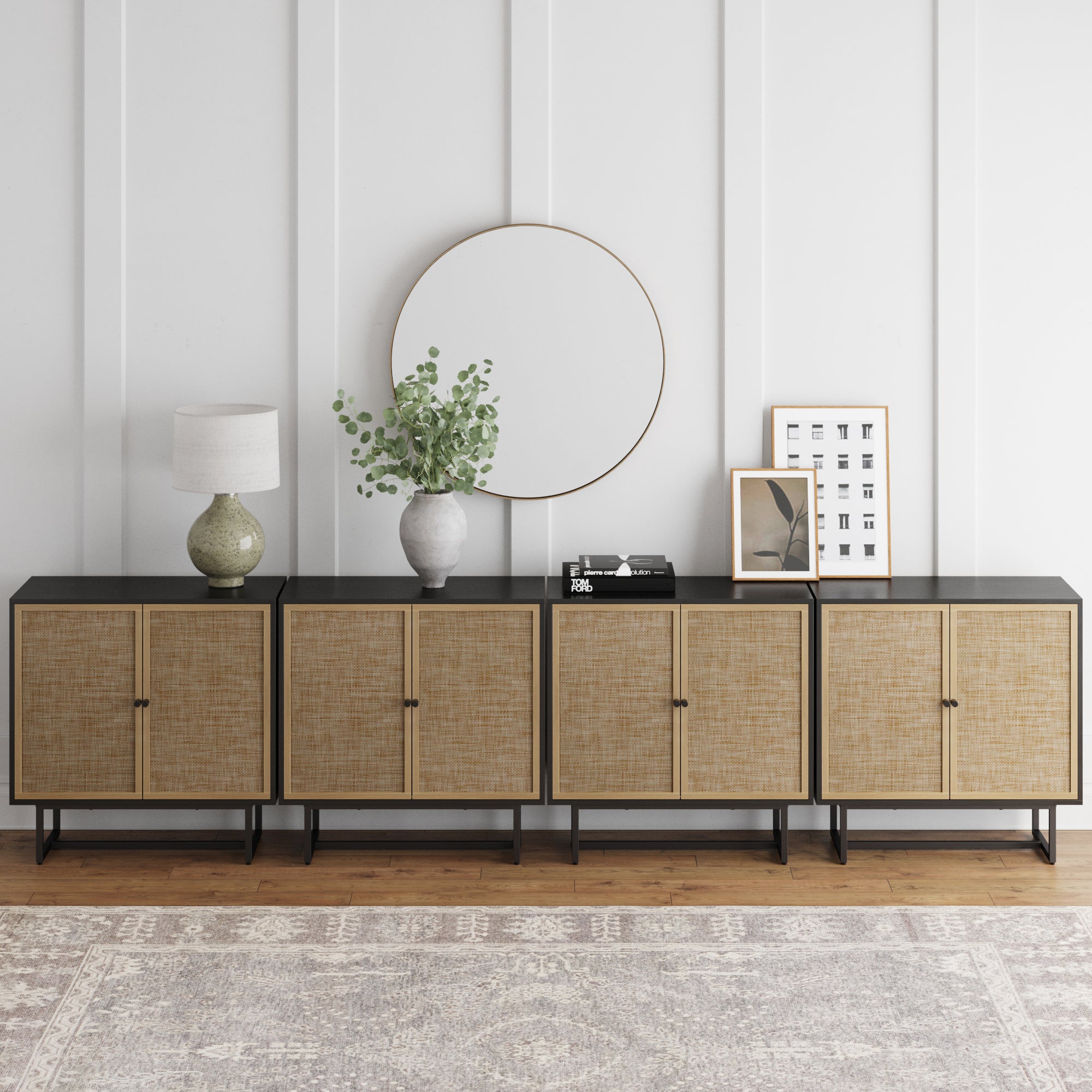 A modern sideboard with four cane-front cabinets sits against a white paneled wall. On top are a round mirror, a lamp, a vase with greenery, books, and framed art. A patterned rug lies on the wooden floor.