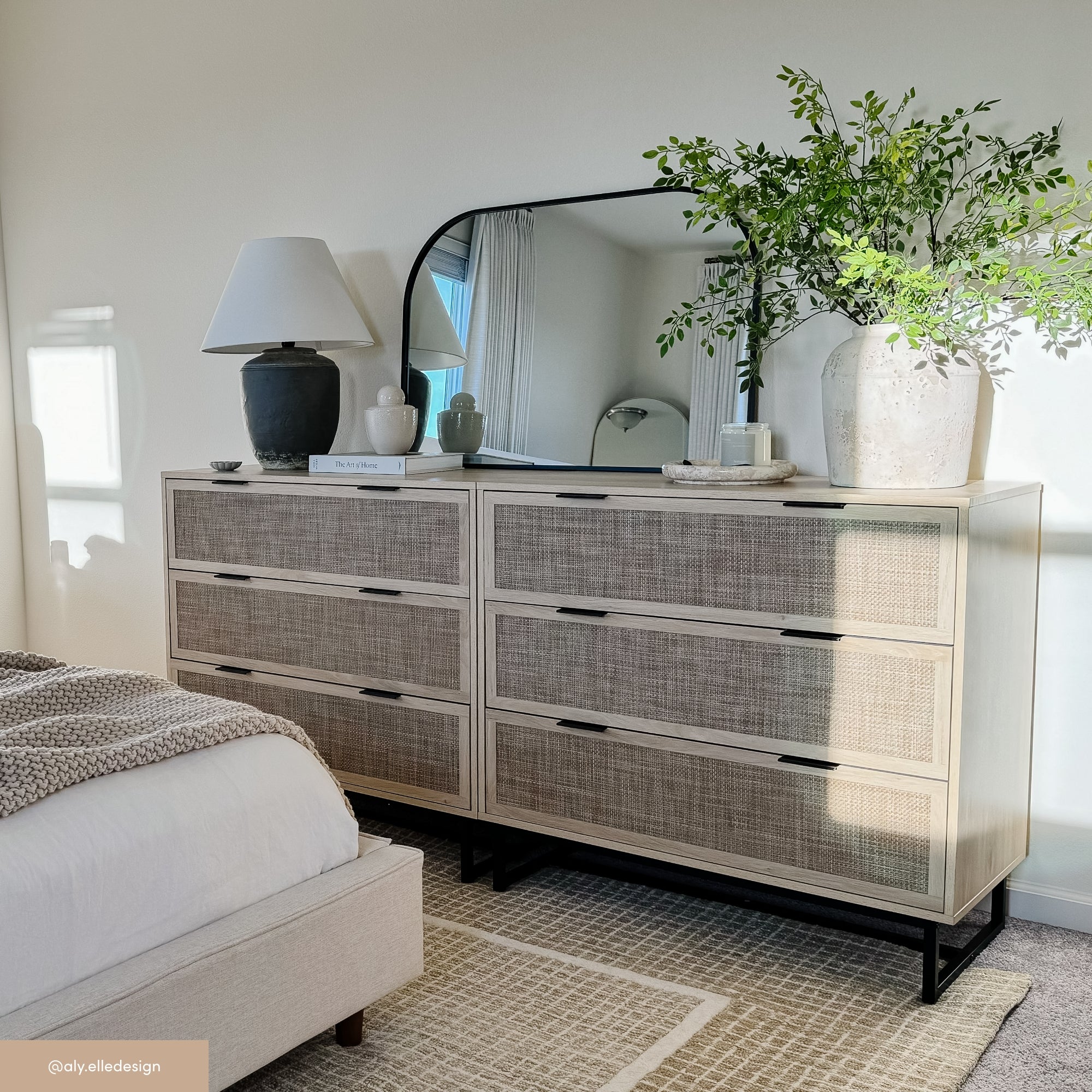 A modern bedroom dresser with six drawers, topped with a black lamp, stacked books, a curved mirror, decorative tray, and a large vase holding leafy branches. Part of a bed and a woven rug are also visible.