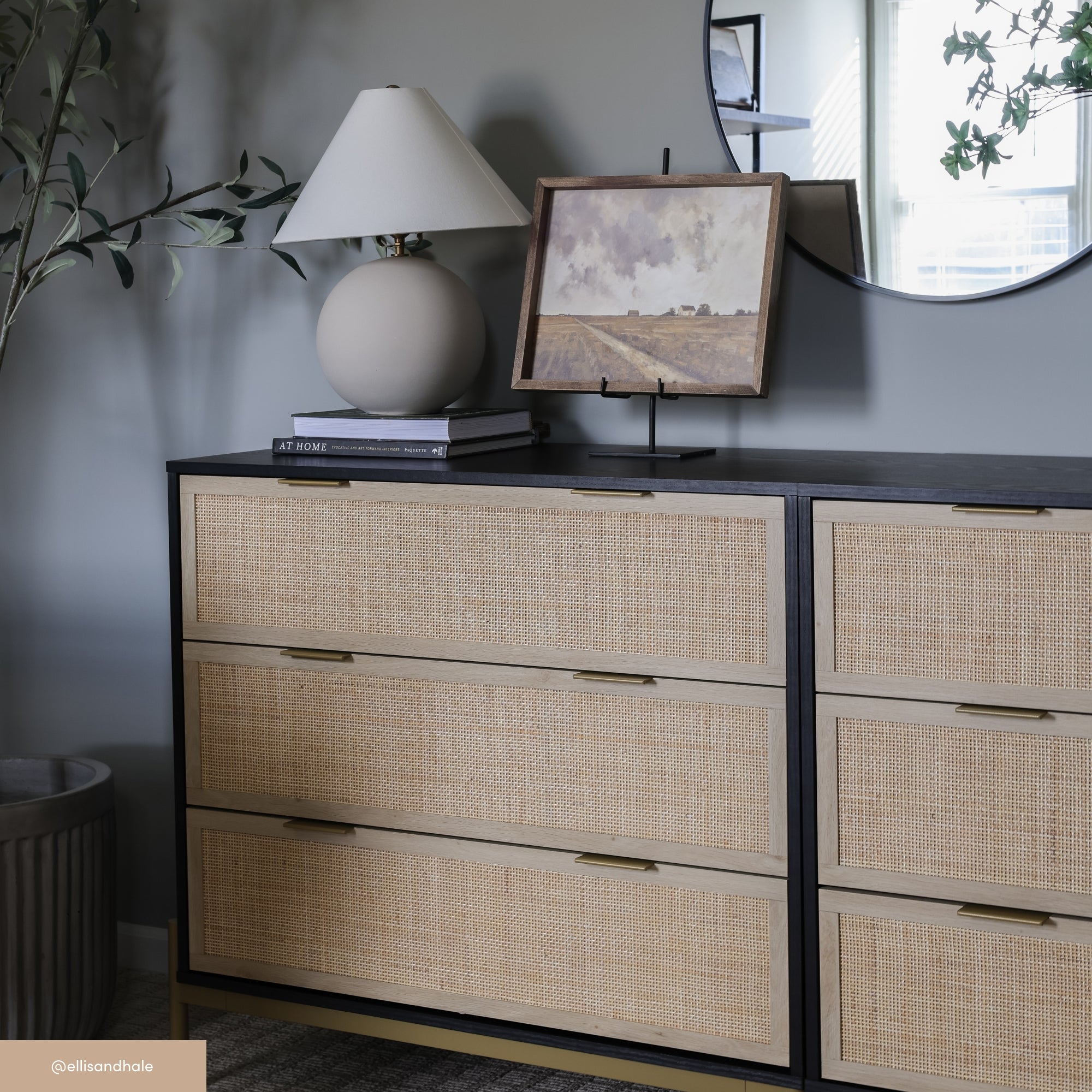 A modern dresser with woven rattan drawers topped with a round lamp and a framed landscape painting, set against a gray wall with a round mirror and a leafy plant nearby.