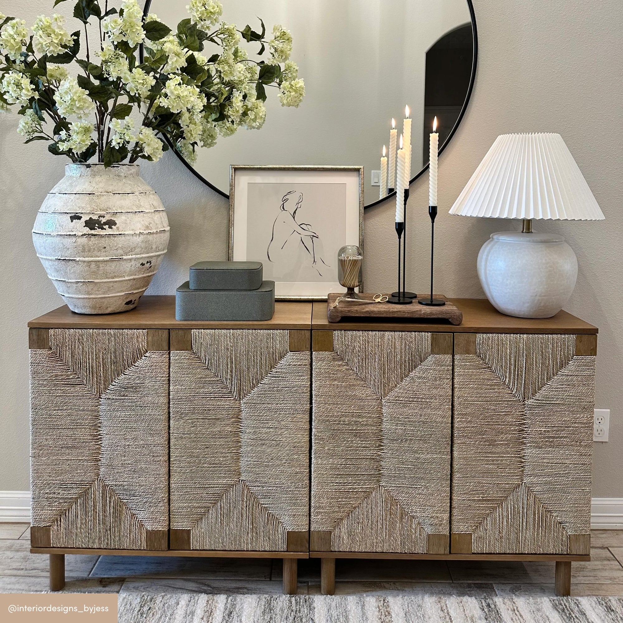 A sideboard with textured doors holds a large vase of white flowers, two green boxes, framed art, candles, and a white table lamp. A round mirror hangs above, reflecting part of the room.
