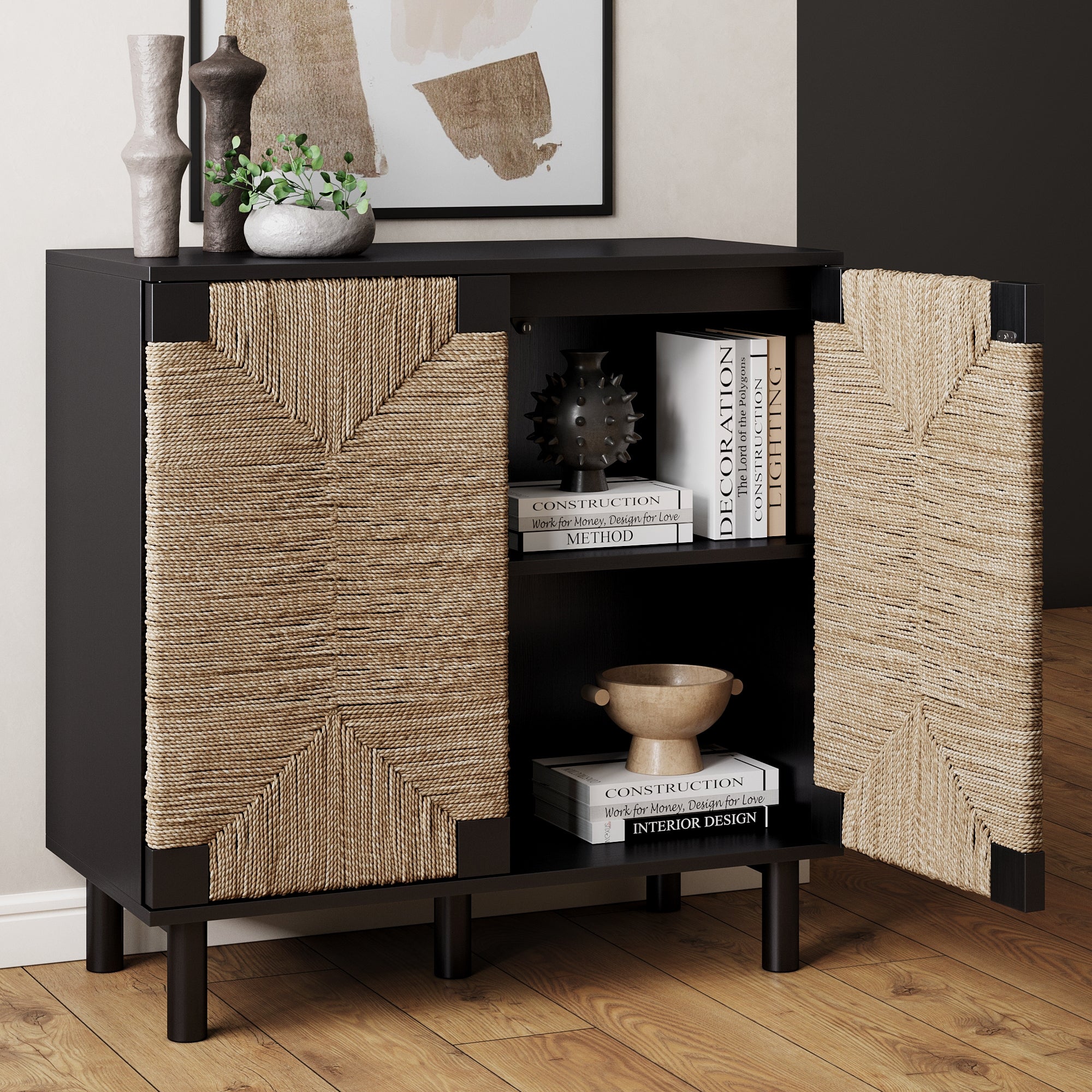 A black wooden cabinet with legs and two open doors featuring a woven rattan design, displaying decorative vases, stacked books, and a bowl on interior shelves; a framed abstract art piece hangs above.