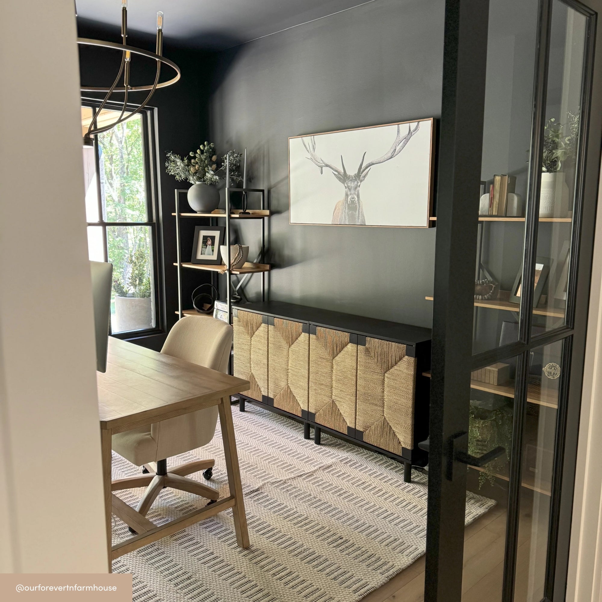 A modern home office with dark walls, a wooden desk and chair, light patterned rug, large sideboard with textured doors, shelves with decor items, a window, and a framed deer artwork on the back wall.