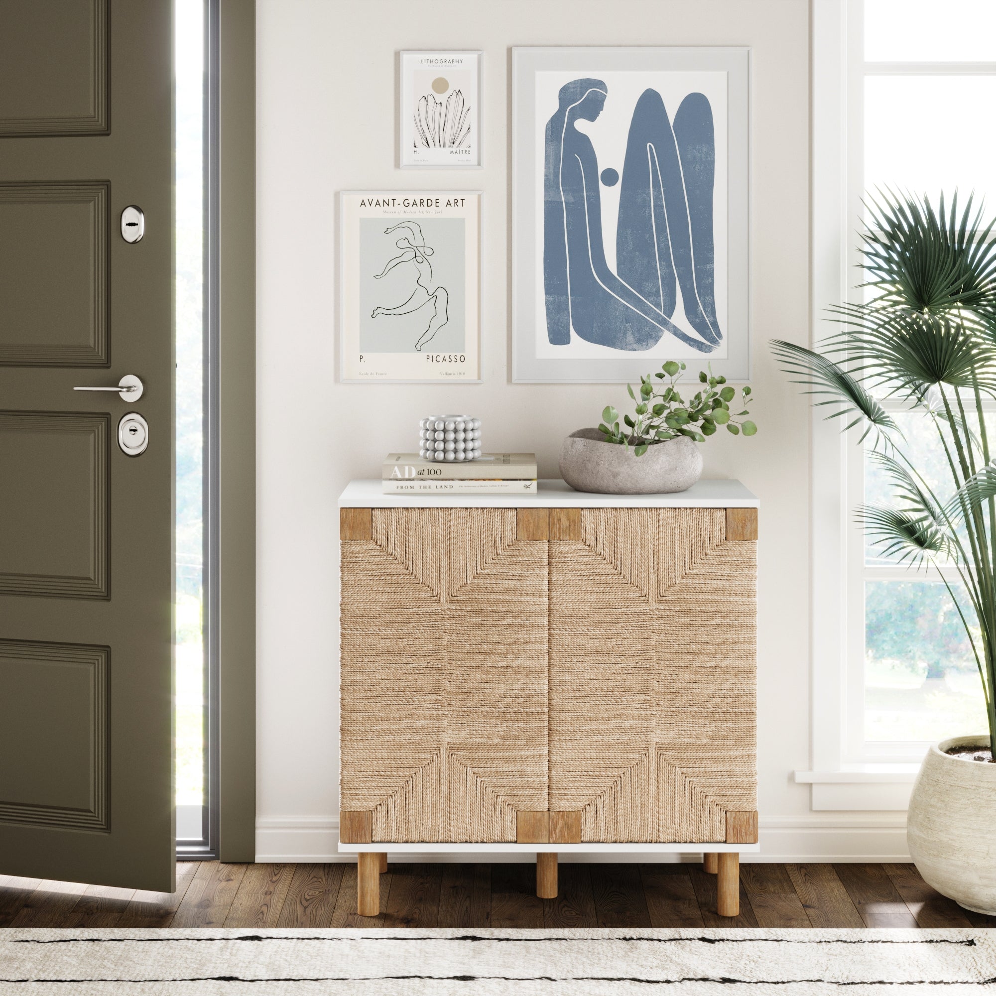 A small modern entryway with a woven cabinet, potted plant, stacked books, and minimalist art prints on a white wall next to a large window and partially open olive green door.