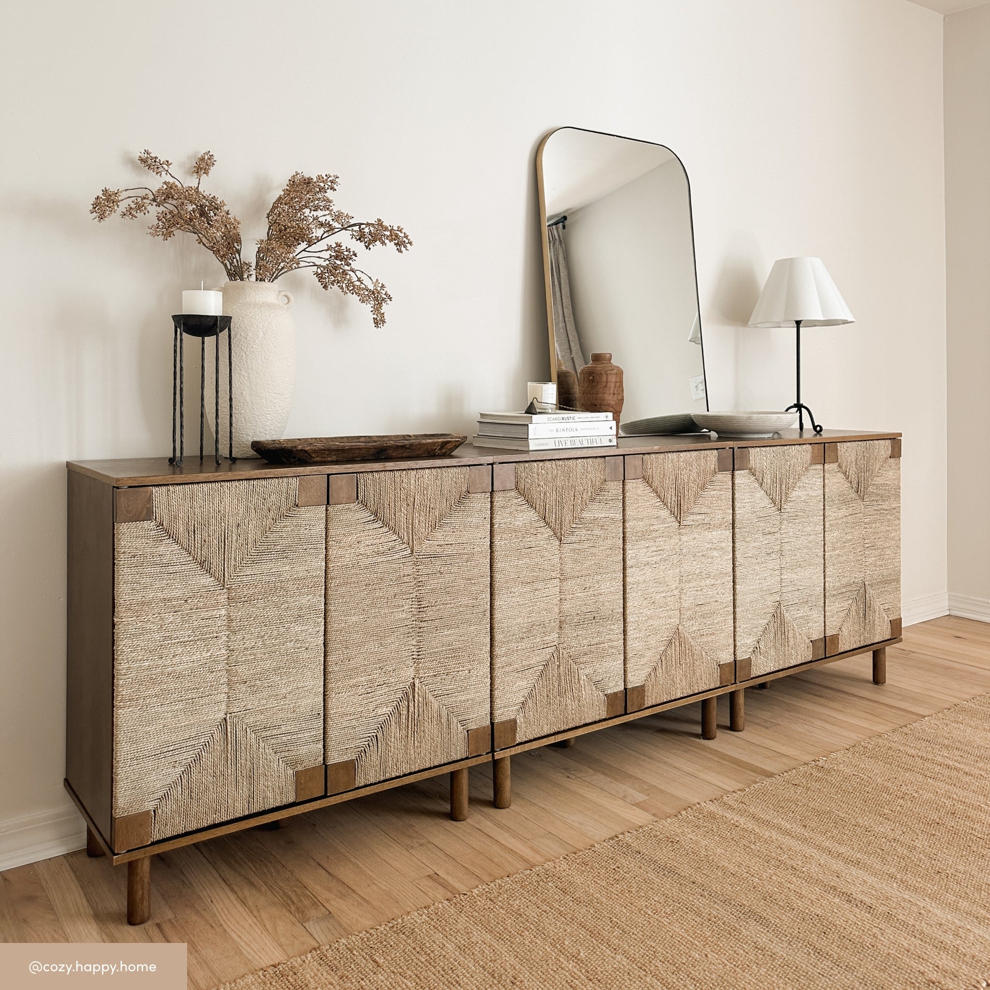 A wooden sideboard with textured, patterned doors stands against a light wall. On top are a large vase with dried flowers, a round mirror, a lamp, books, and decorative objects. A woven rug covers the floor.