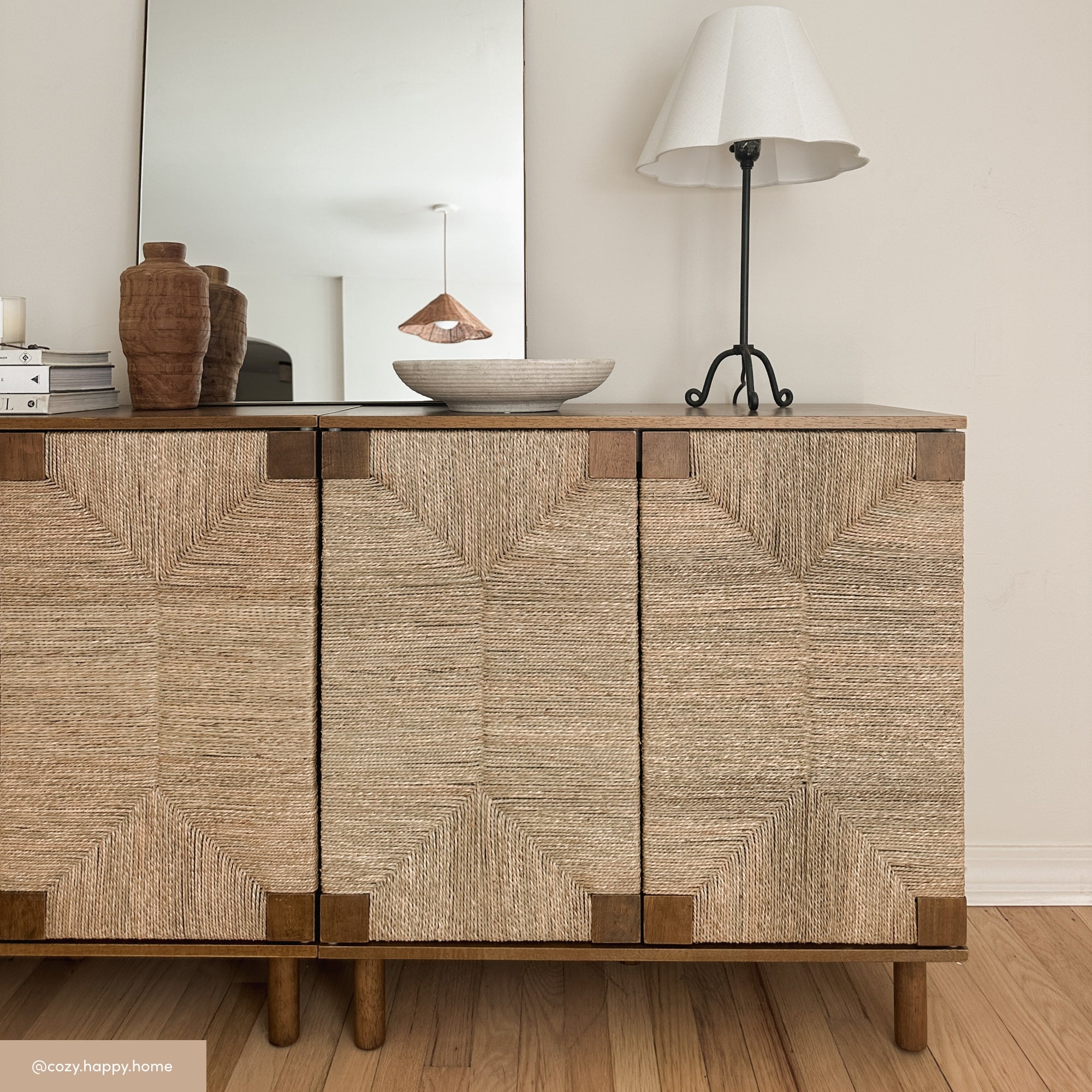 A woven cabinet sits on a wooden floor, topped with a white table lamp, brown vases, and a bowl. A large mirror behind the cabinet reflects a pendant light and a white wall.