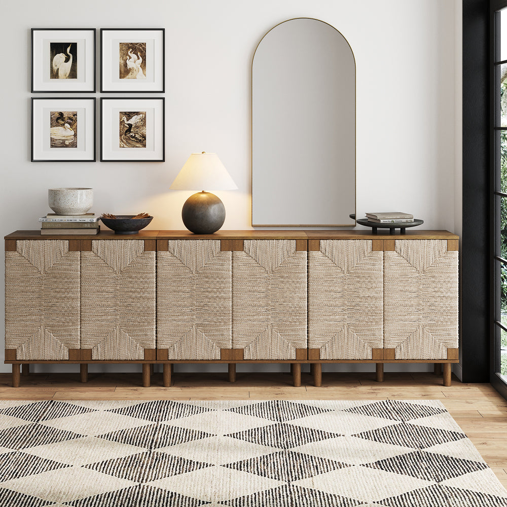 A wide wooden sideboard with woven fiber door panels sits below an arched wall mirror, a table lamp, and four framed prints of birds.