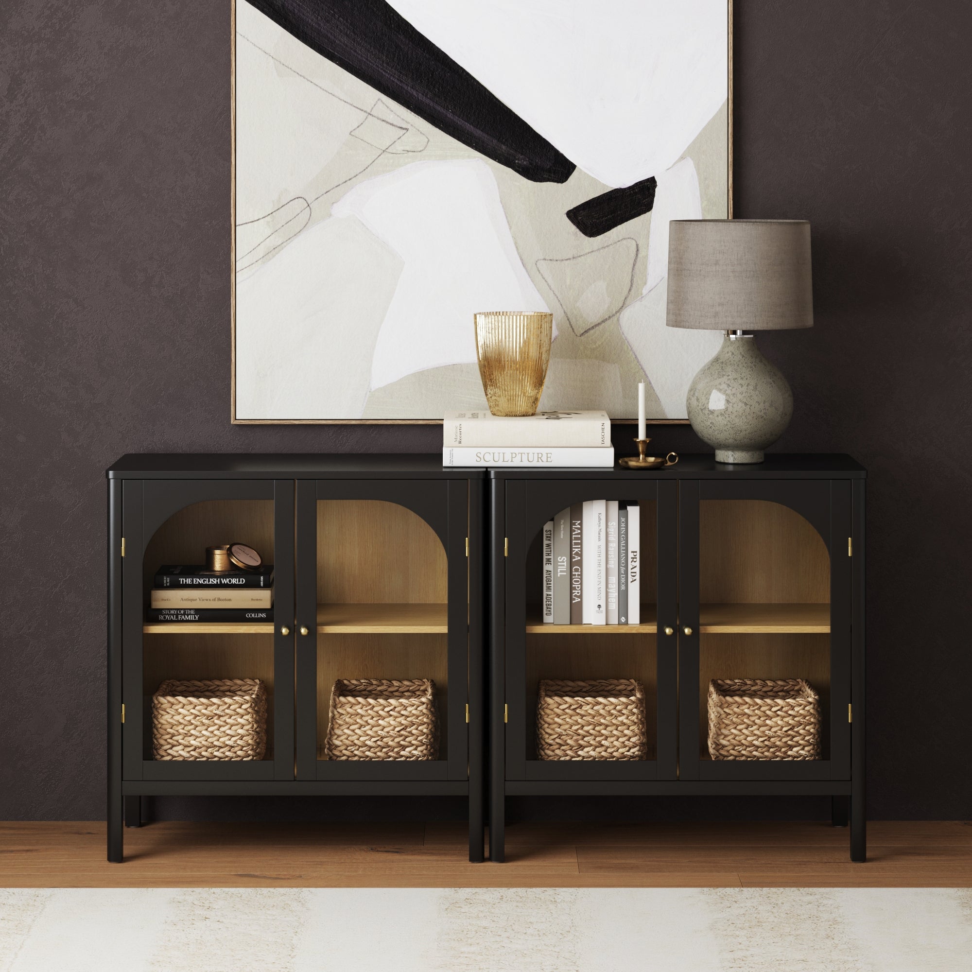 A black sideboard with glass doors holds woven baskets and books, topped with decor items like a lamp, vase, and glass bowl, set against a dark wall with abstract artwork above.