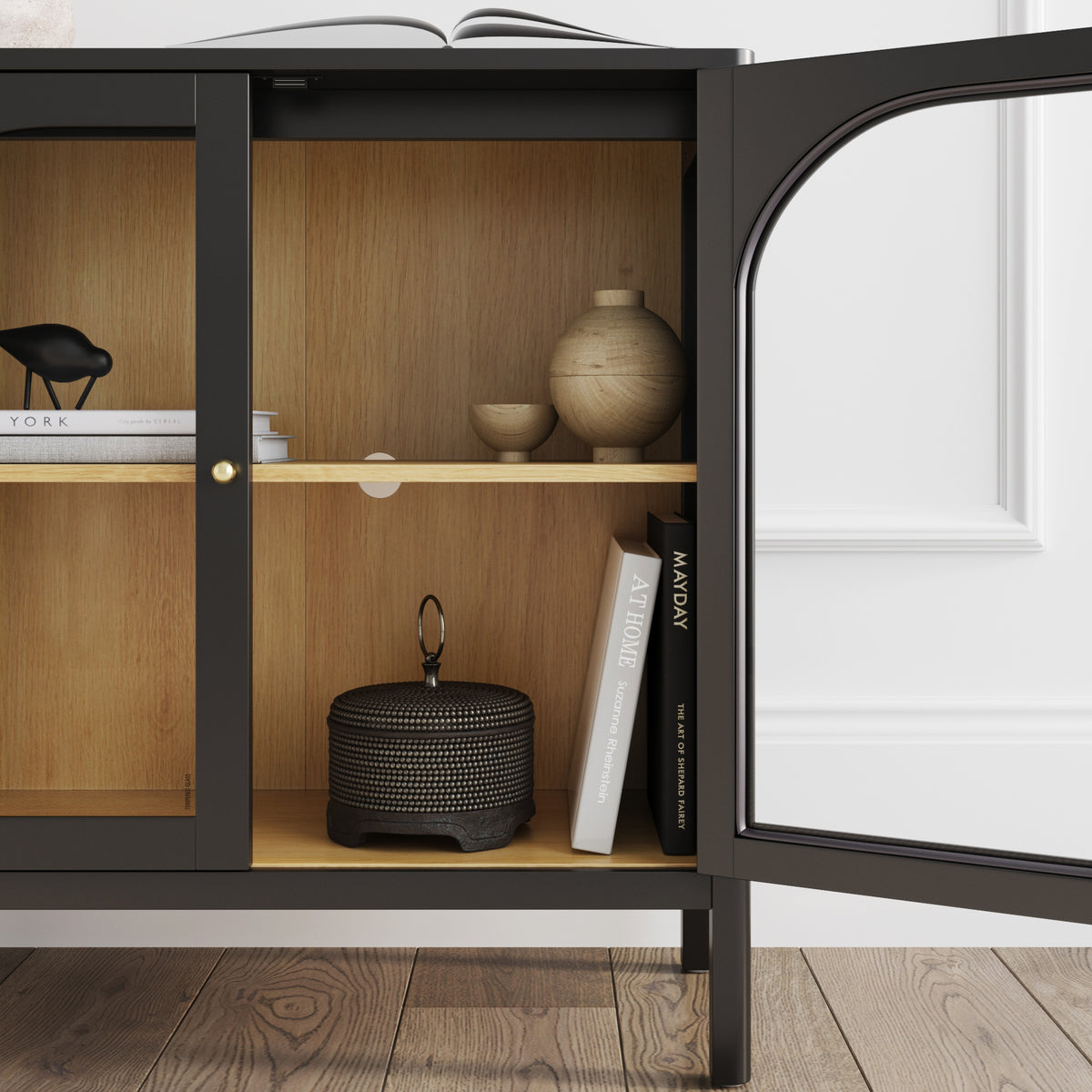 A black Nathan James Curio Cabinet Credenza with Glass Door Black, featuring a wooden interior, an arched glass door, and shelves displaying decorative bowls and books.