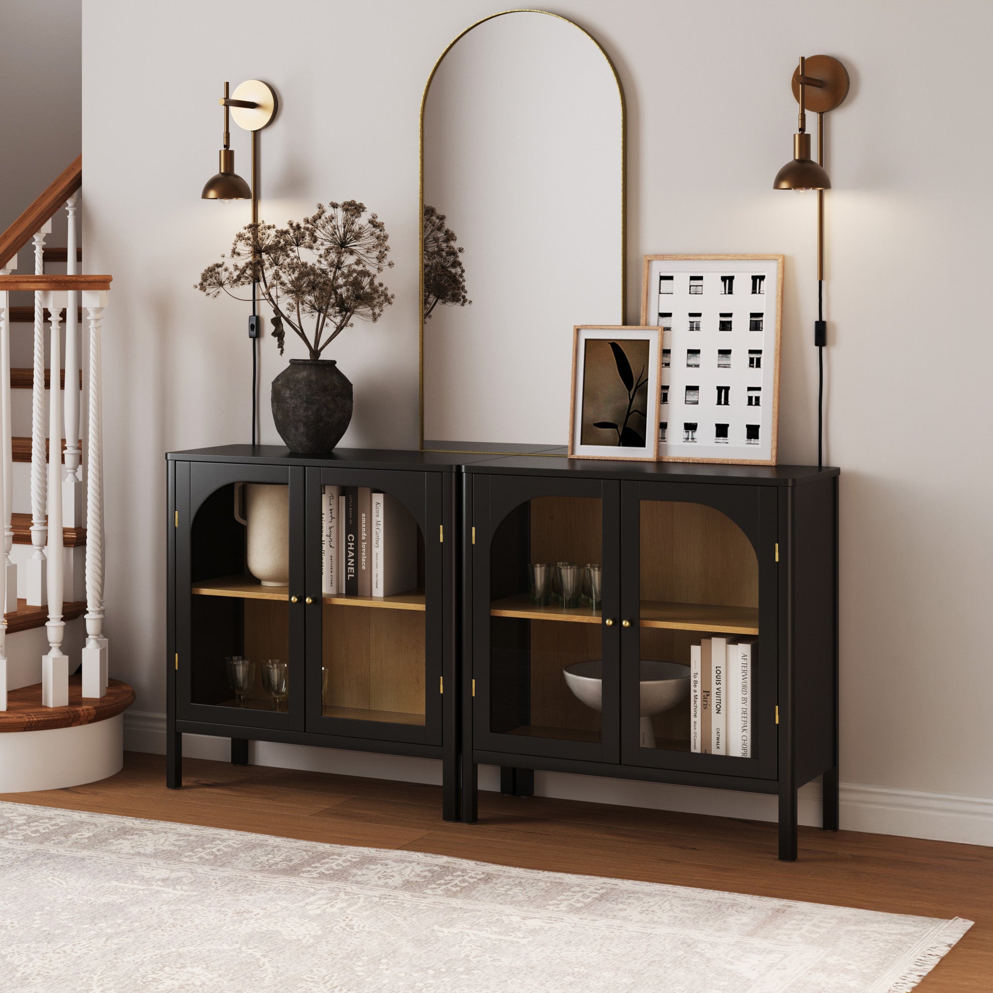 A modern hallway features two black cabinets with glass doors, a decorative vase with branches, framed art, a large arched mirror, and wall-mounted sconces beneath a staircase. The area is warmly lit and neatly arranged.