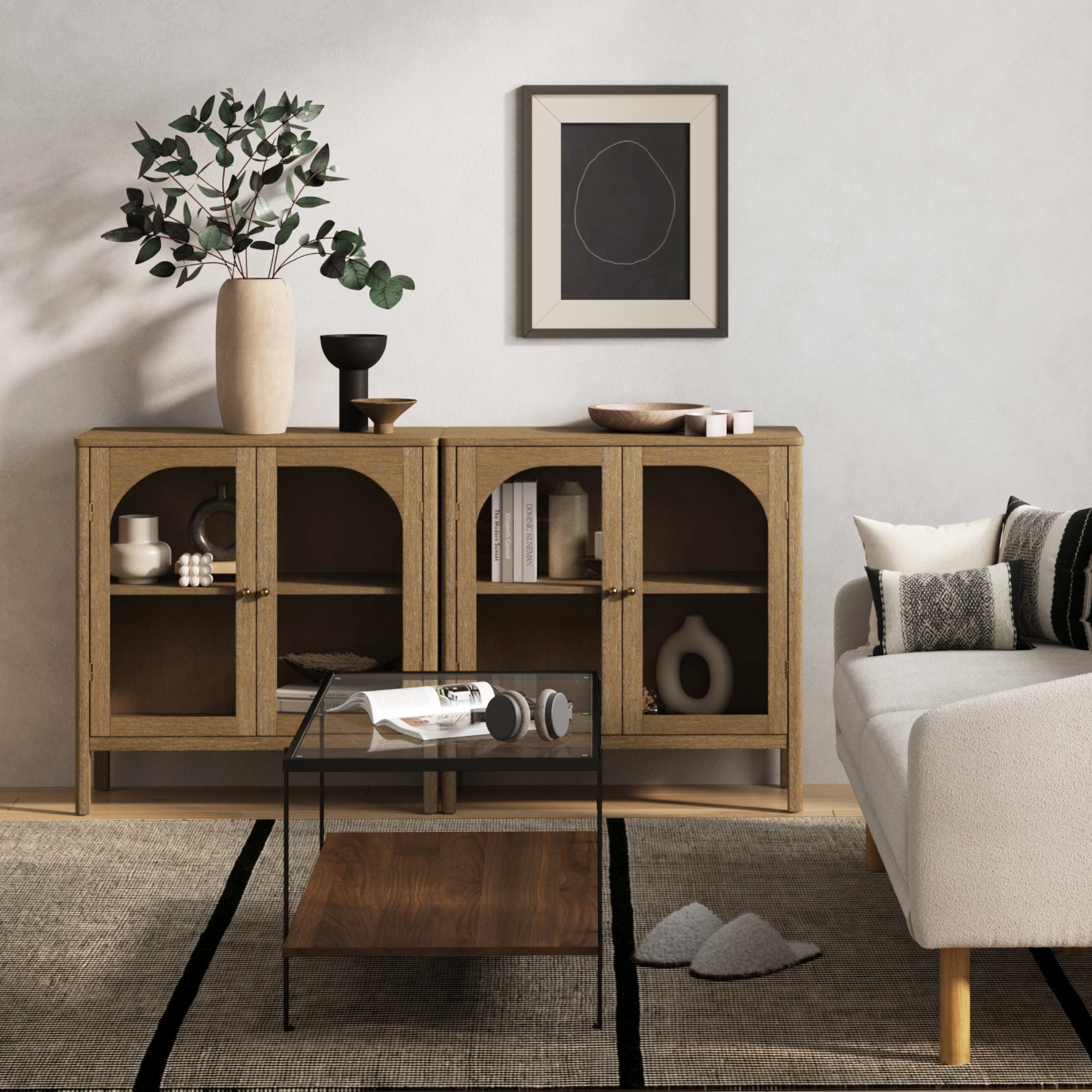 A modern living room with a wooden sideboard featuring glass doors, a large ceramic vase with greenery, decorative objects, a framed abstract print, a white sofa with cushions, and a rug with a small coffee table.