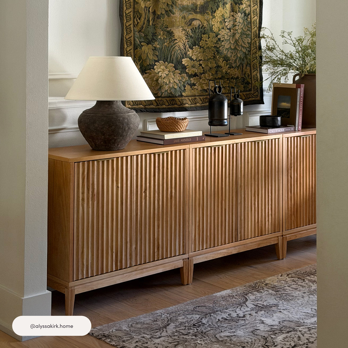 A set of three Nathan James Mid-Century Fluted Storage Cabinet Credenza Warm Pine units with fluted doors arranged in a row, topped with a table lamp, books, and decorative objects.