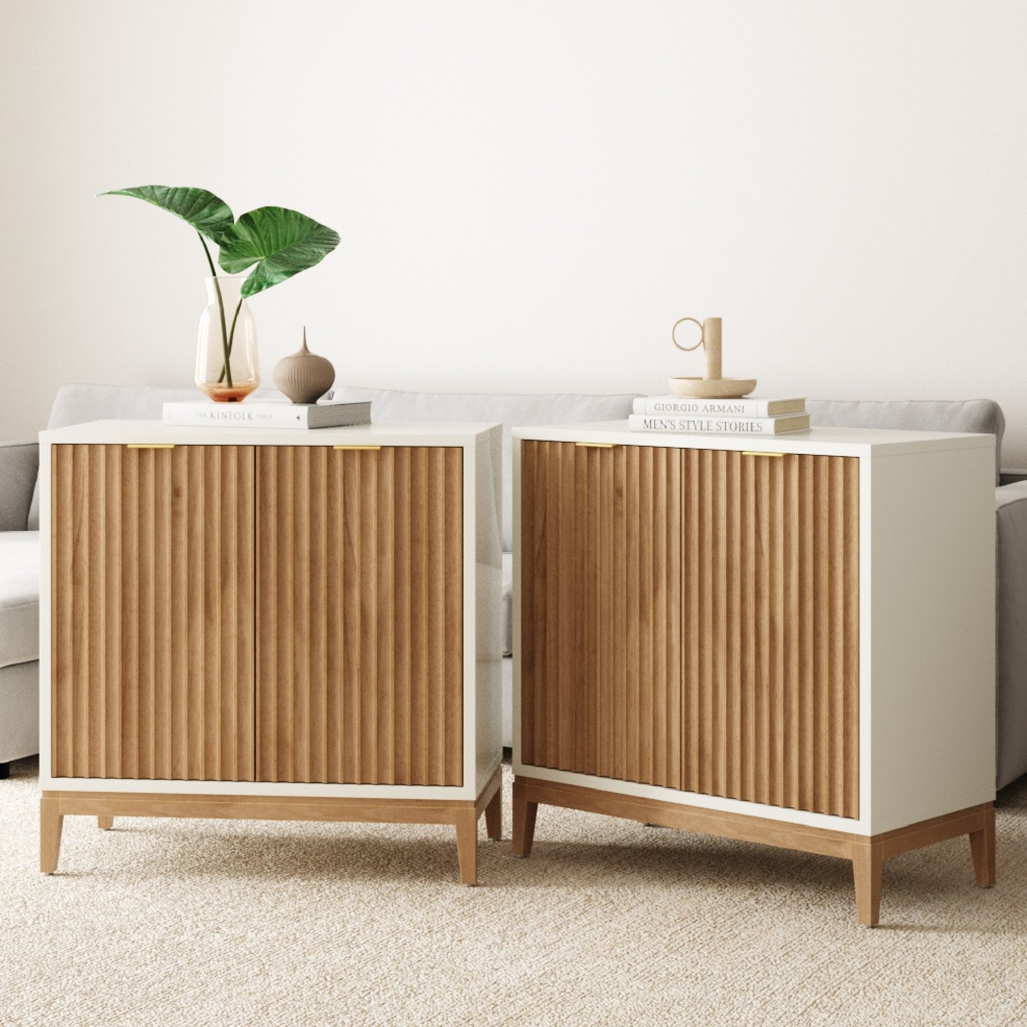 Two modern wooden cabinets with vertical grooves and white frames sit side by side in a living room. Decorative items, such as books, vases, and a plant, are placed on top. A light-colored sofa is in the background.