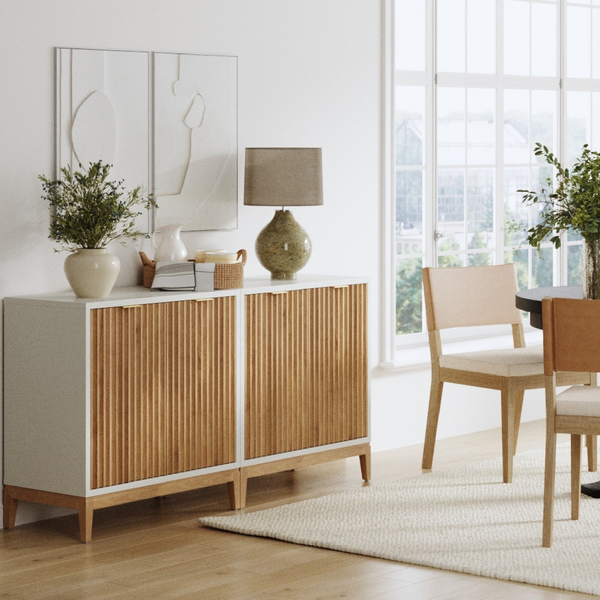 A modern dining room with light wood furniture, a white and wood sideboard decorated with plants, ceramics, books, and a lamp, a dining table with chairs, large windows, and neutral decor.