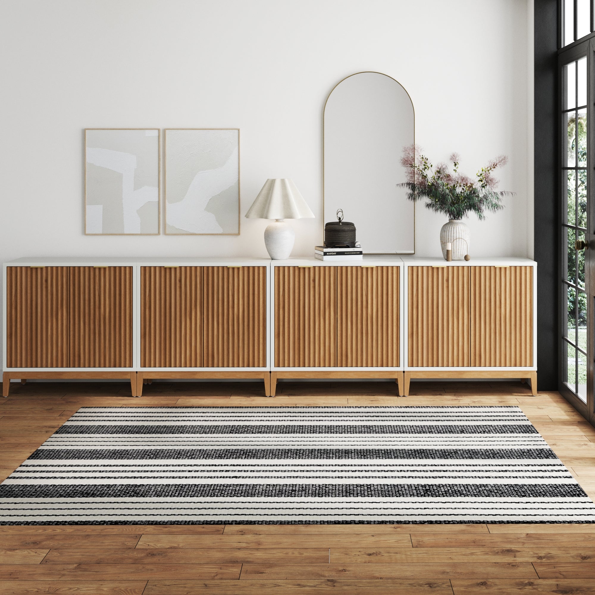A modern living room with a wooden sideboard, two abstract artworks, a lamp, a mirror, and a vase with flowers. A black and white striped rug covers the wooden floor, and large windows let in natural light.