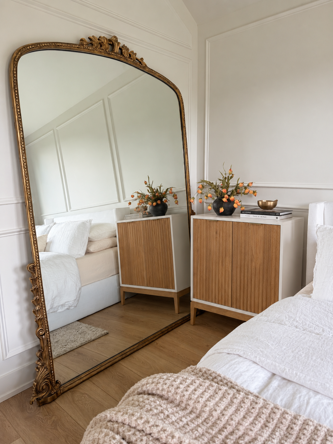 An arched, ornate gold floor mirror reflecting a bedroom with a bed and a wooden cabinet topped with a vase of orange flowers.