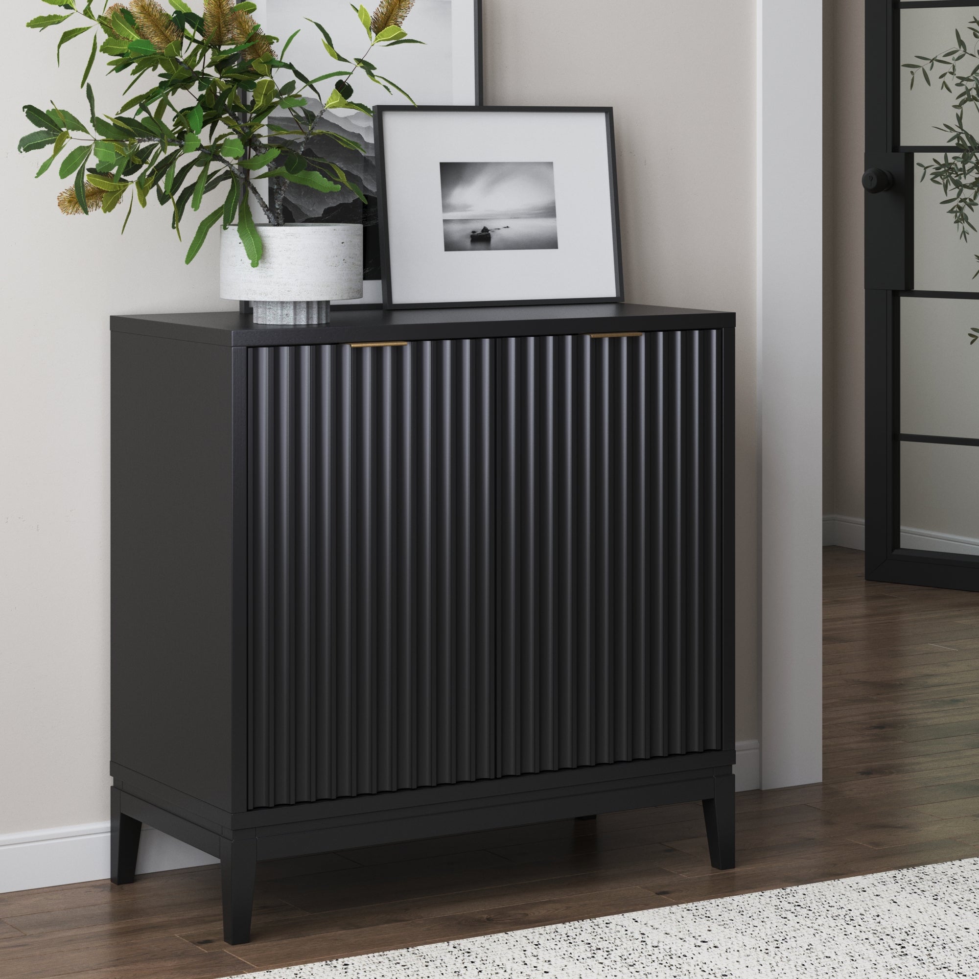 A modern black cabinet with ribbed doors stands against a beige wall. On top, there’s a potted plant and a framed black-and-white photo. Part of a rug and a glass door are visible nearby.