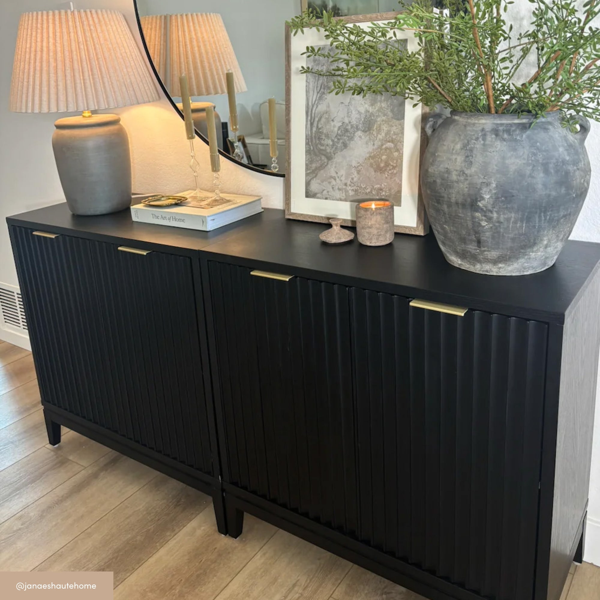 A black sideboard with gold handles is decorated with a large gray vase holding greenery, a smaller gray vase, a candle, two stacked books, and a beige lamp with a pleated shade, set against a mirror on a wood floor.