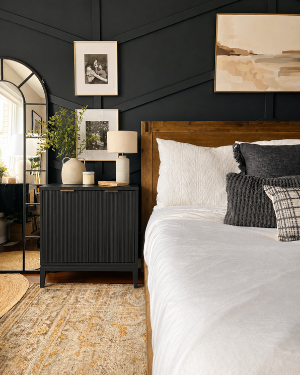 A bed with a wood frame and white bedding sits next to a black ribbed nightstand topped with a lamp and a plant, against a black paneled wall decorated with framed art.