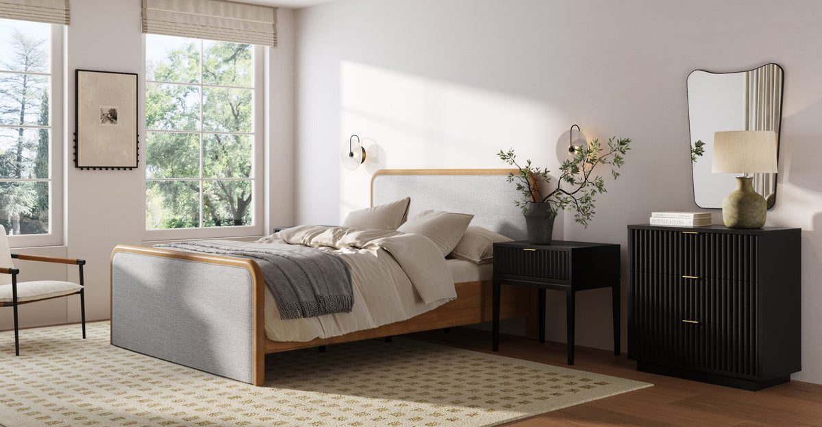 A bedroom with a light wood and grey fabric bed, a black nightstand, and a matching black dresser with an irregular-shaped wall mirror.