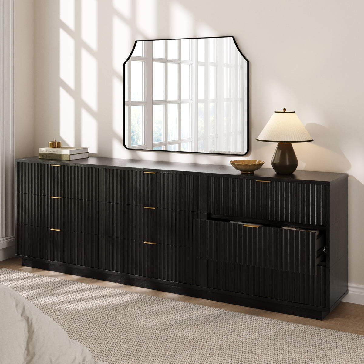 Long black dresser with vertical fluting, gold handles, and one open drawer, topped with a lamp and a mirror on the wall.